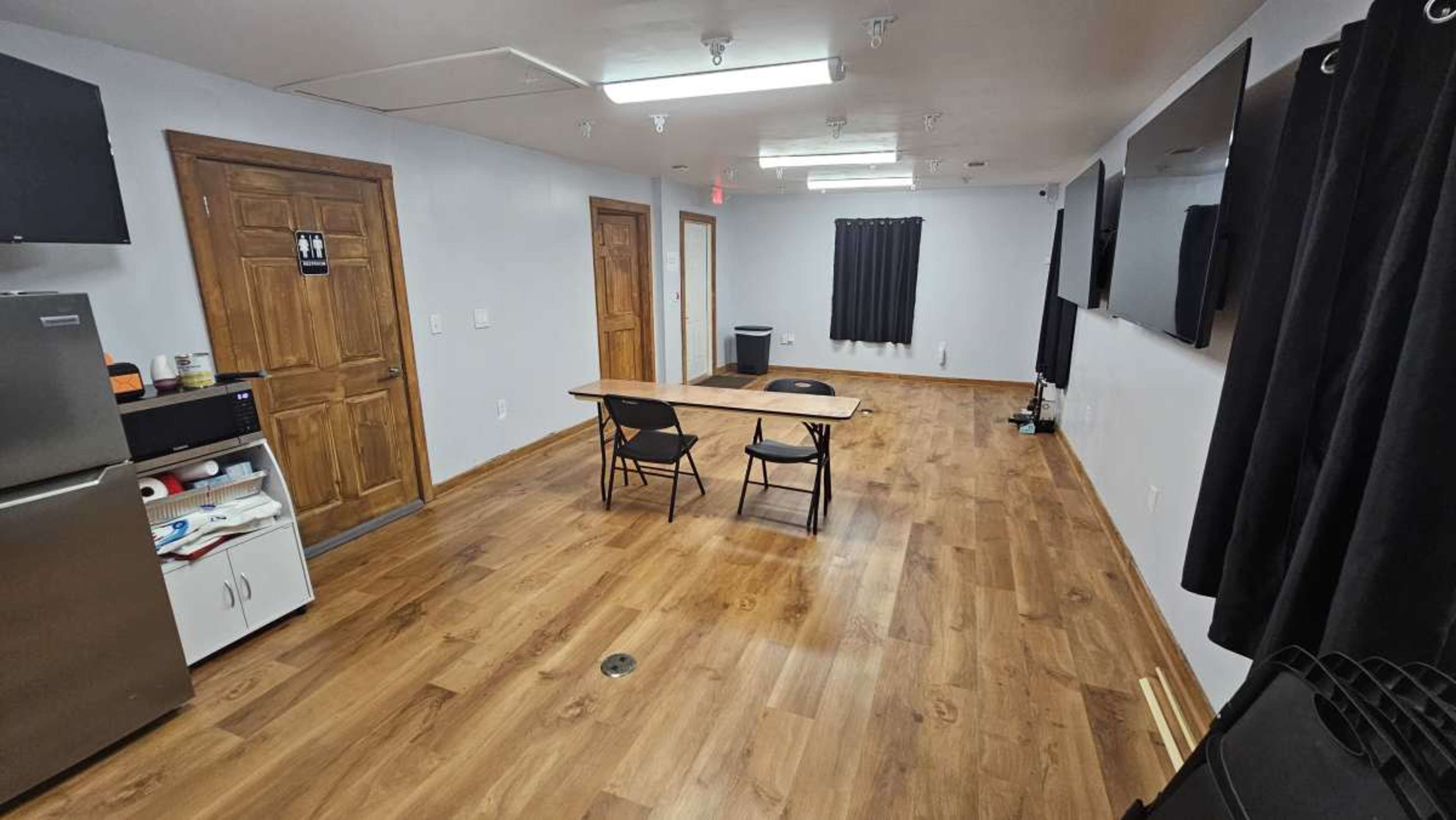 The image shows an empty room with a wooden floor, a table with two chairs, a kitchenette area, and multiple wall-mounted screens.