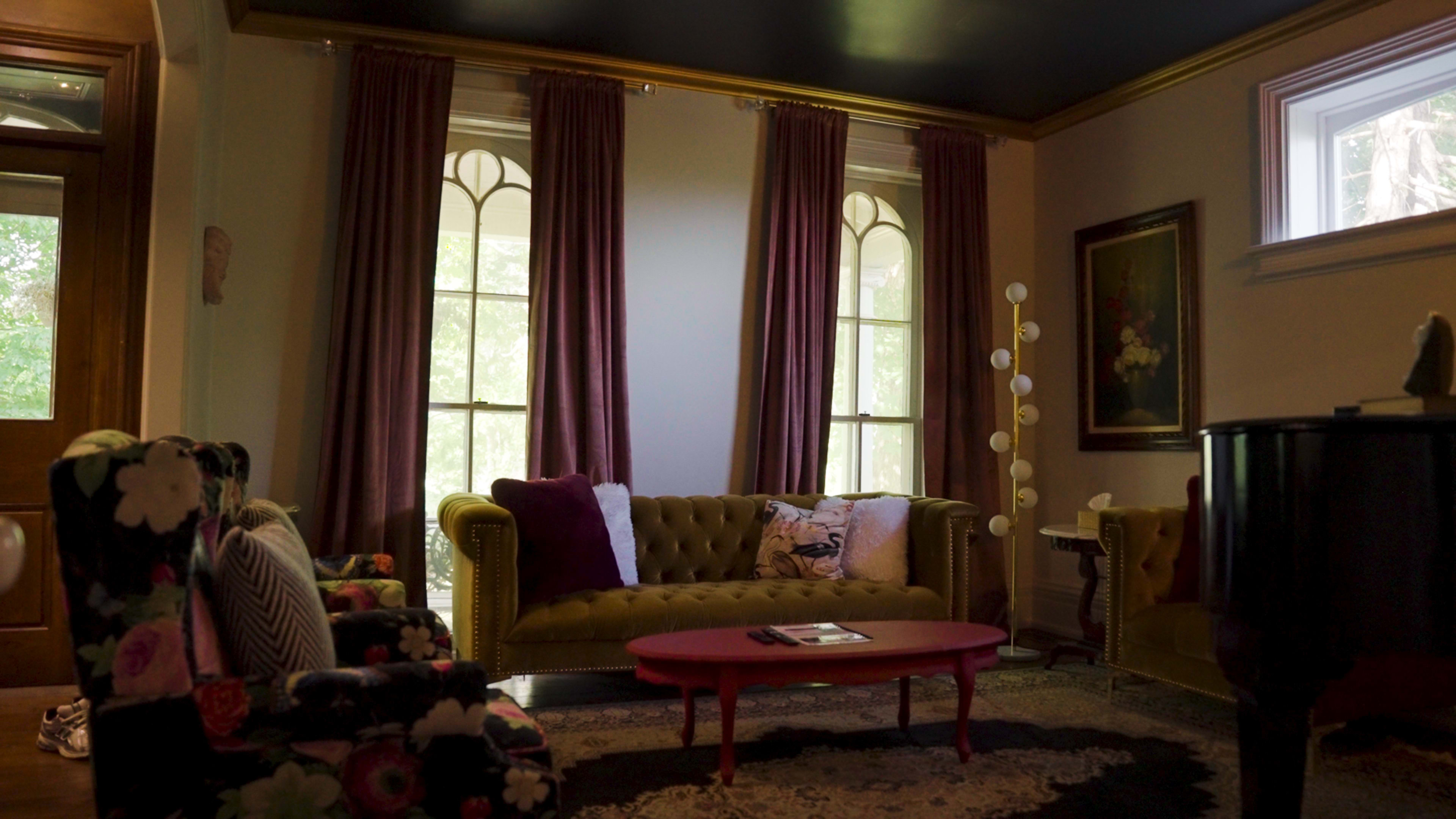 The living room features a green tufted sofa, a pink coffee table, and floral patterned armchairs, with large windows dressed in burgundy curtains.