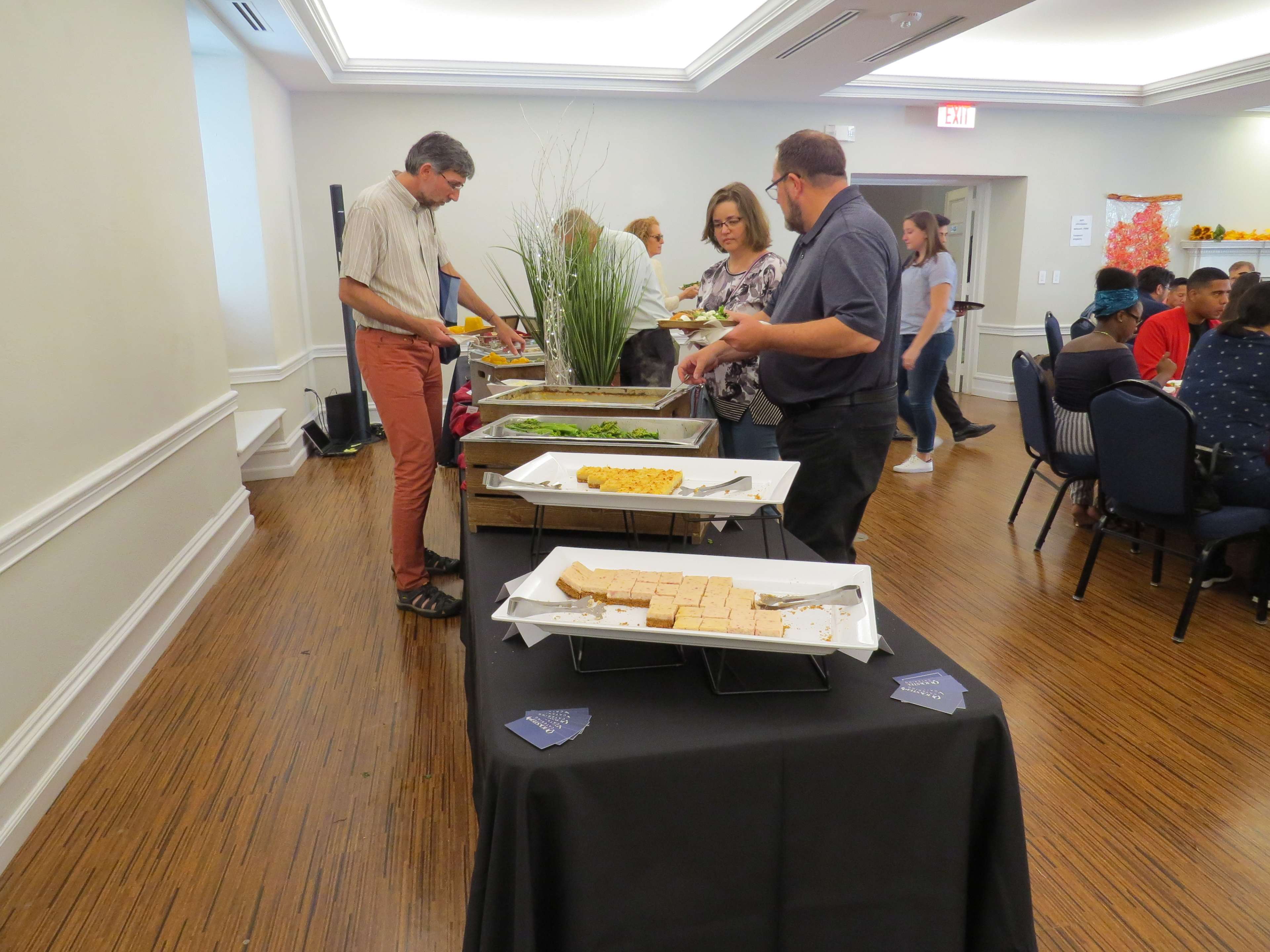 People are serving themselves food from a buffet table in a well-lit room with a gathering of seated attendees in the background.
