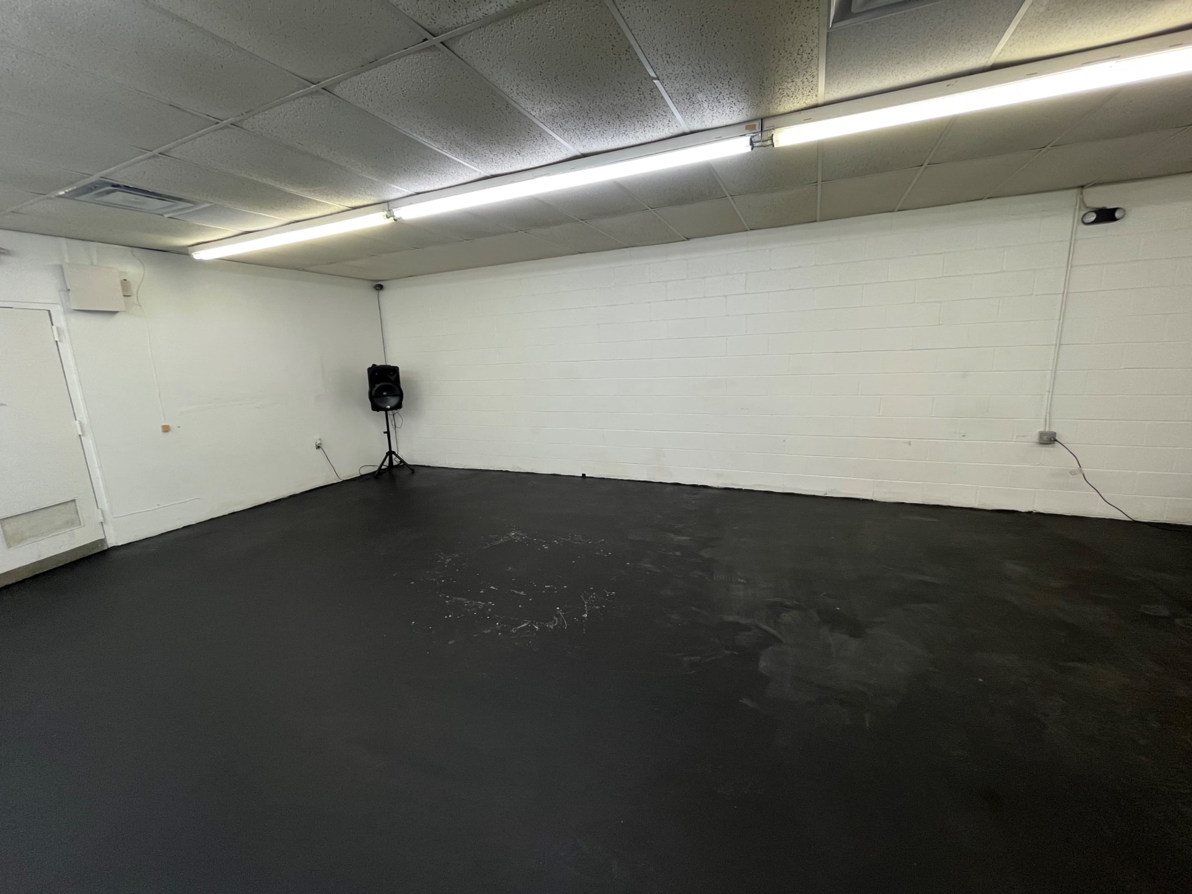 An empty room with a black floor and white walls, featuring a speaker in the corner and overhead fluorescent lighting.
