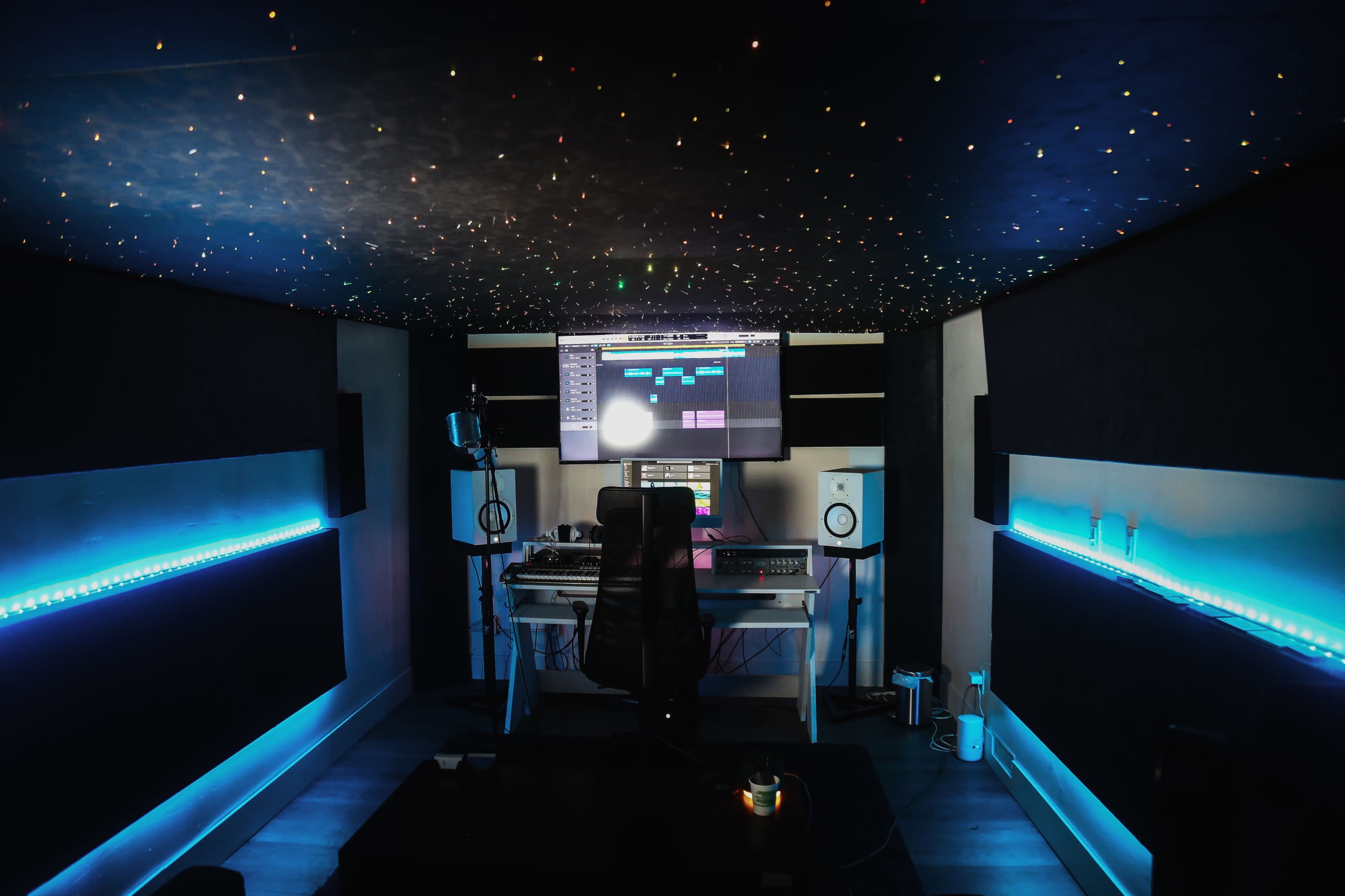 A music production studio features a computer setup with a large monitor, microphone, and illuminated walls.