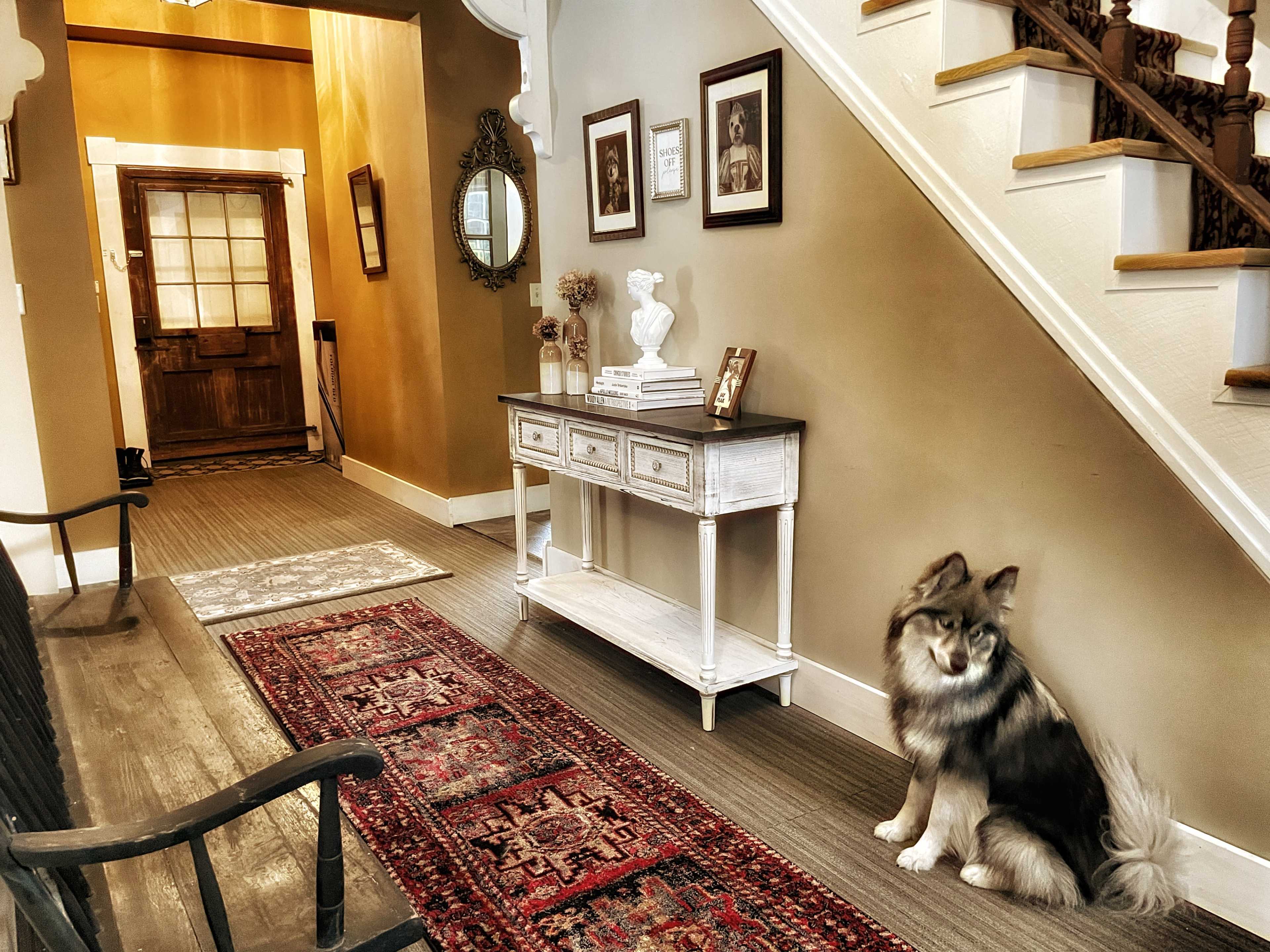 A hallway features a console table against the wall and a dog sitting on a rug, with a staircase leading to the upper floor in the background.