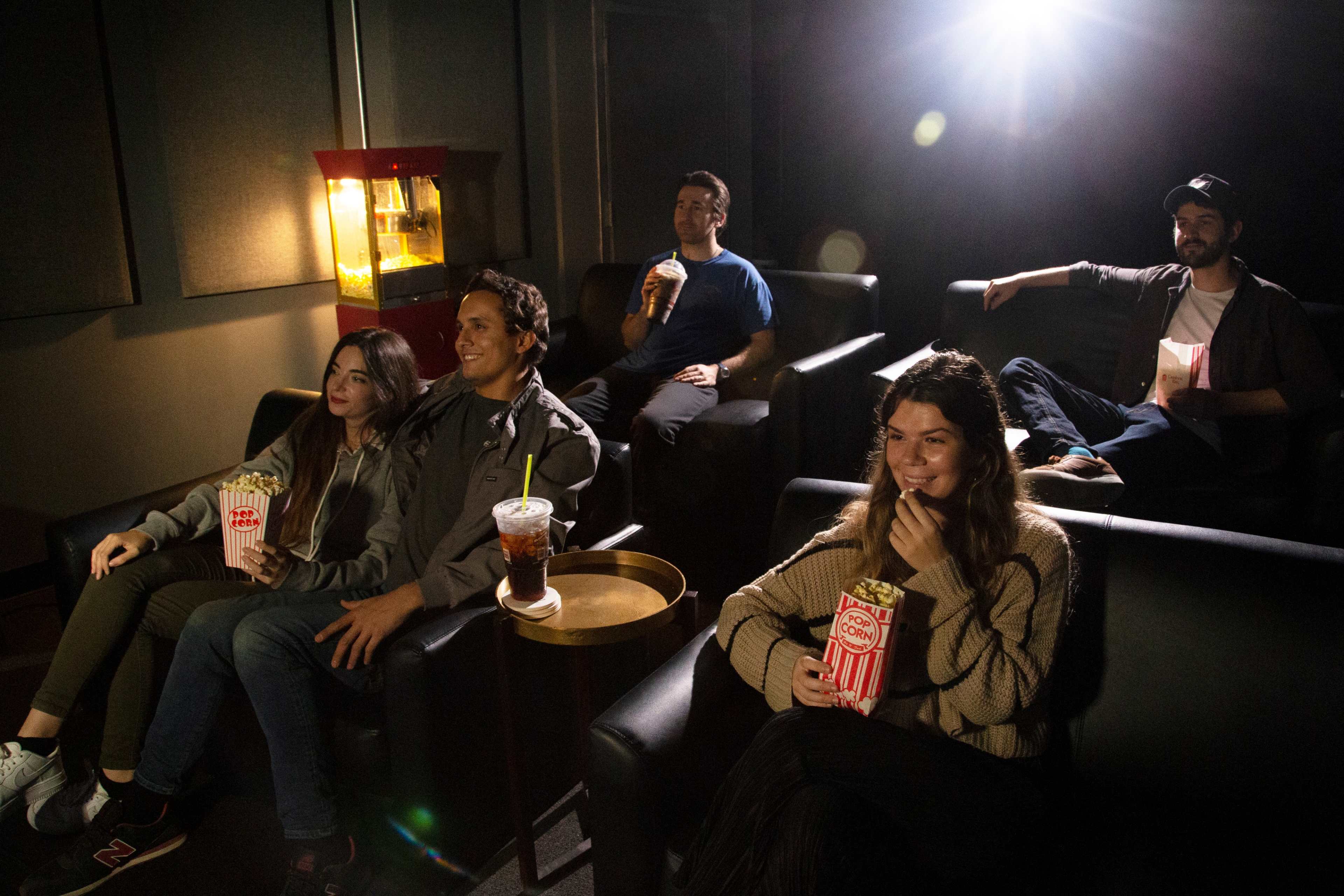 A group of five people sits in a movie theater, with some holding popcorn and drinks, while a projector casts light in the background.
