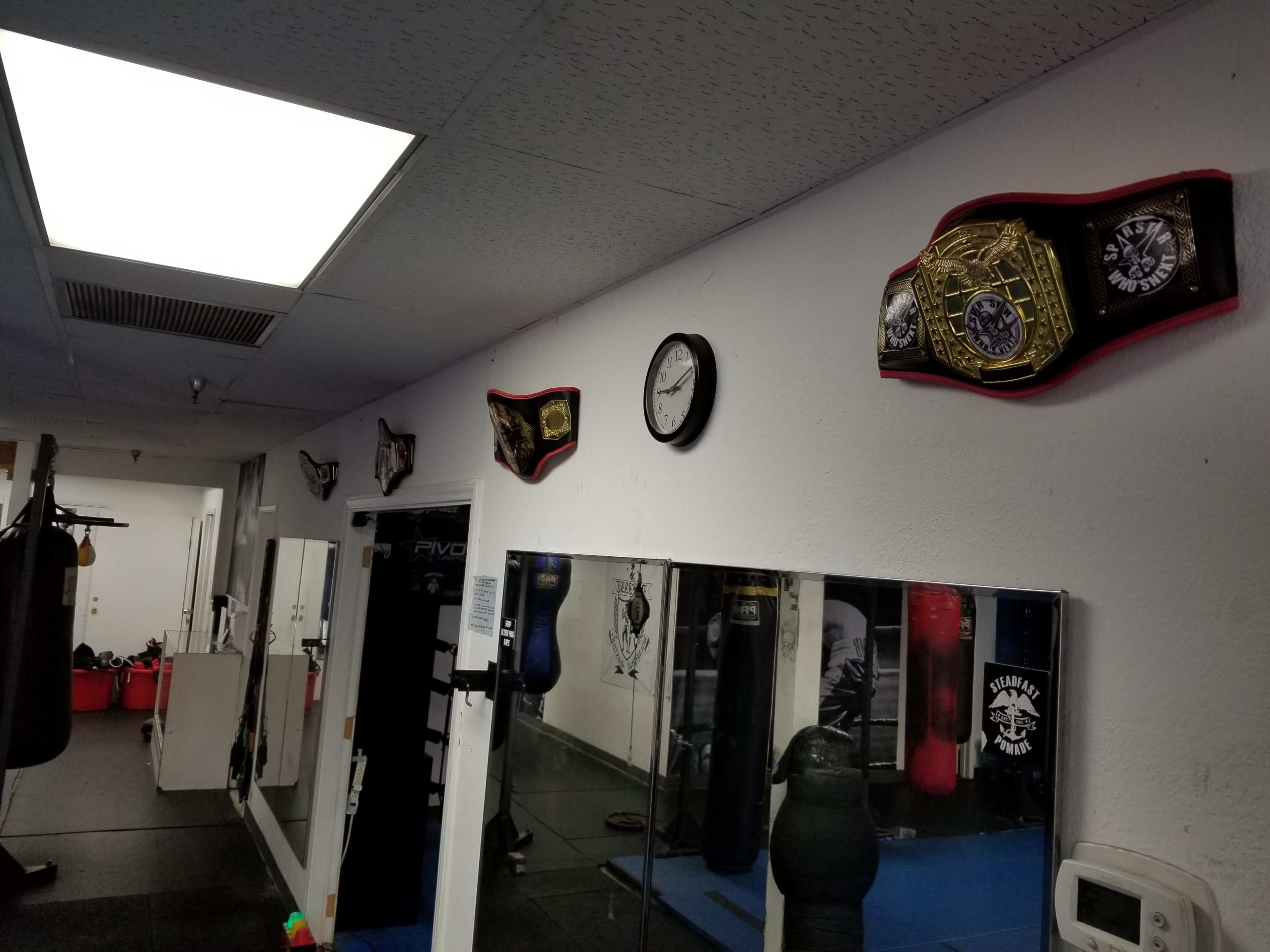 The image shows a gym interior with championship belts mounted on the wall, a clock, and several punching bags reflected in a mirror.