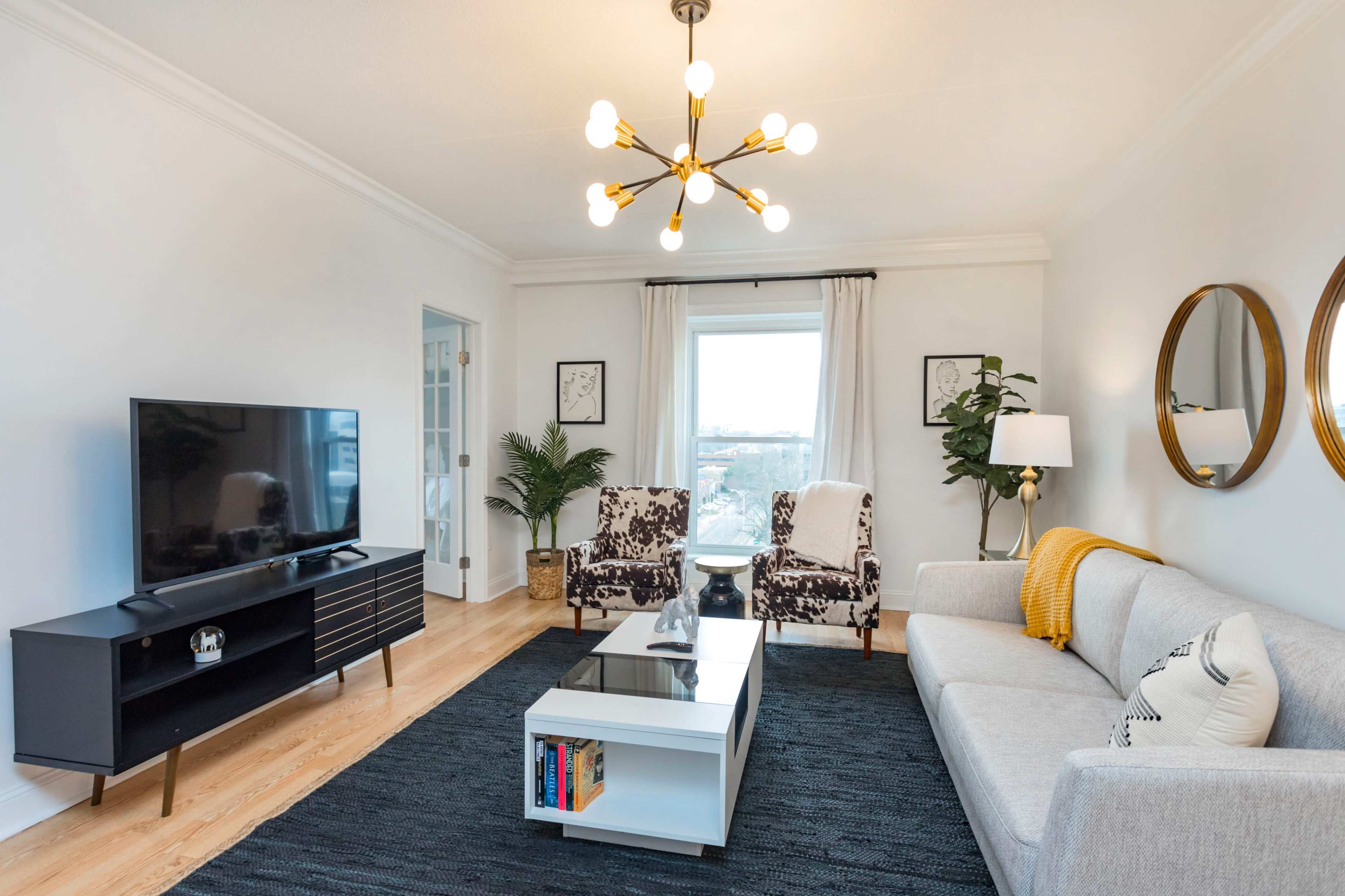 A modern living room features a gray sofa, a black television unit, a coffee table, and two patterned chairs, with a large window providing natural light.