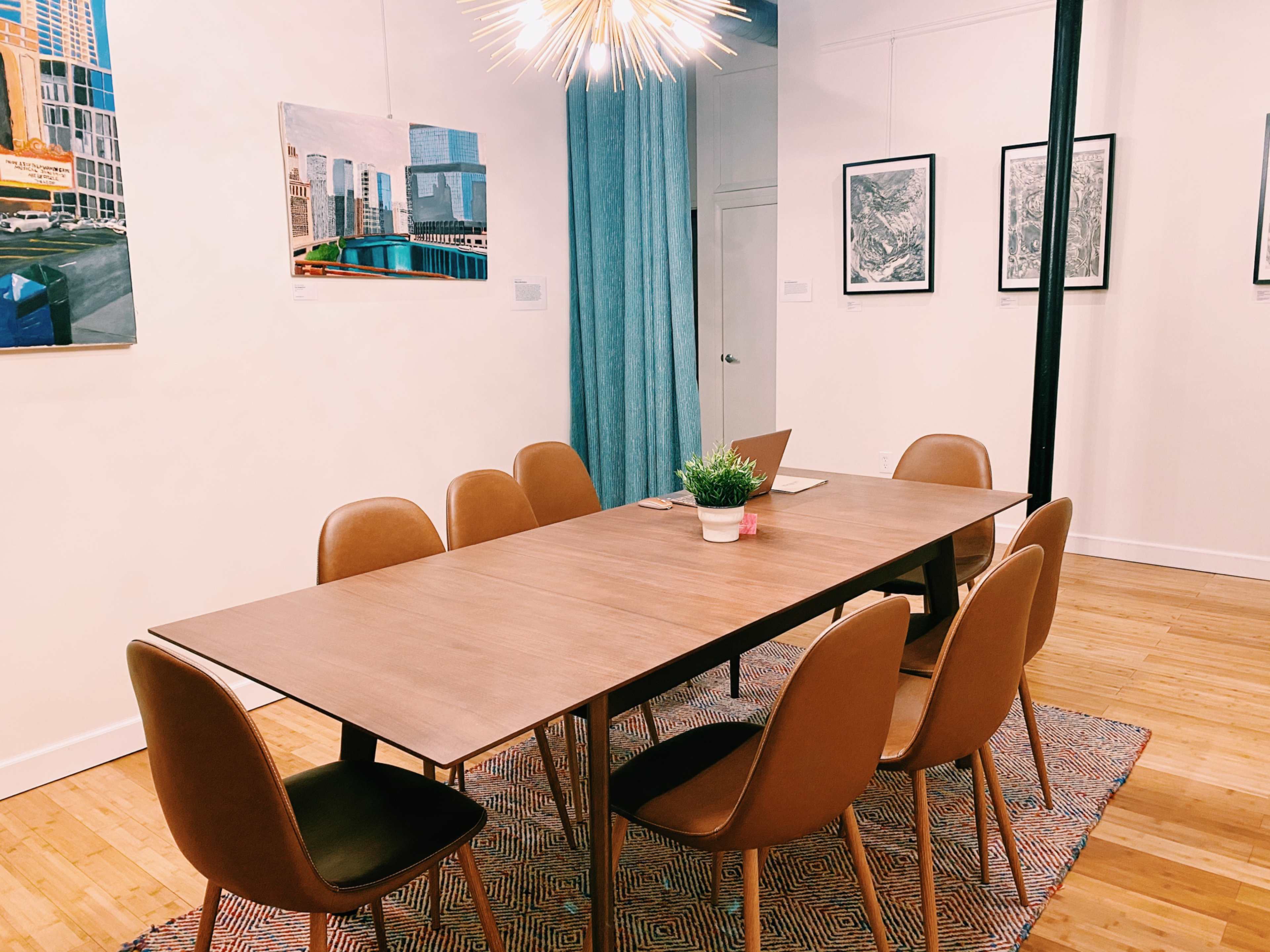 A long wooden table surrounded by brown chairs is set in a room with framed artwork on the walls and a potted plant in the center.
