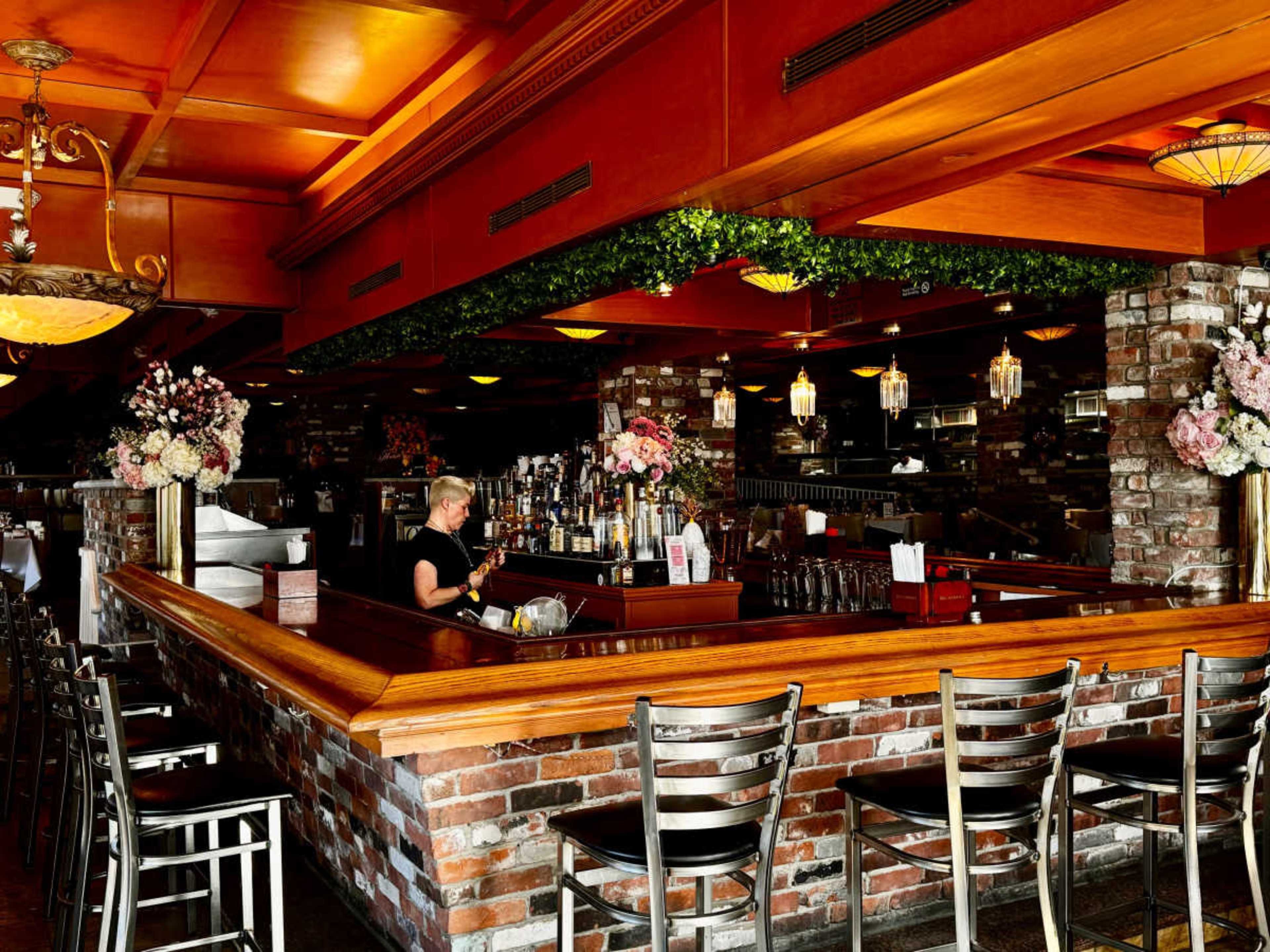 The image shows a bar area with a wooden counter, brick walls, and floral decorations in a restaurant setting.