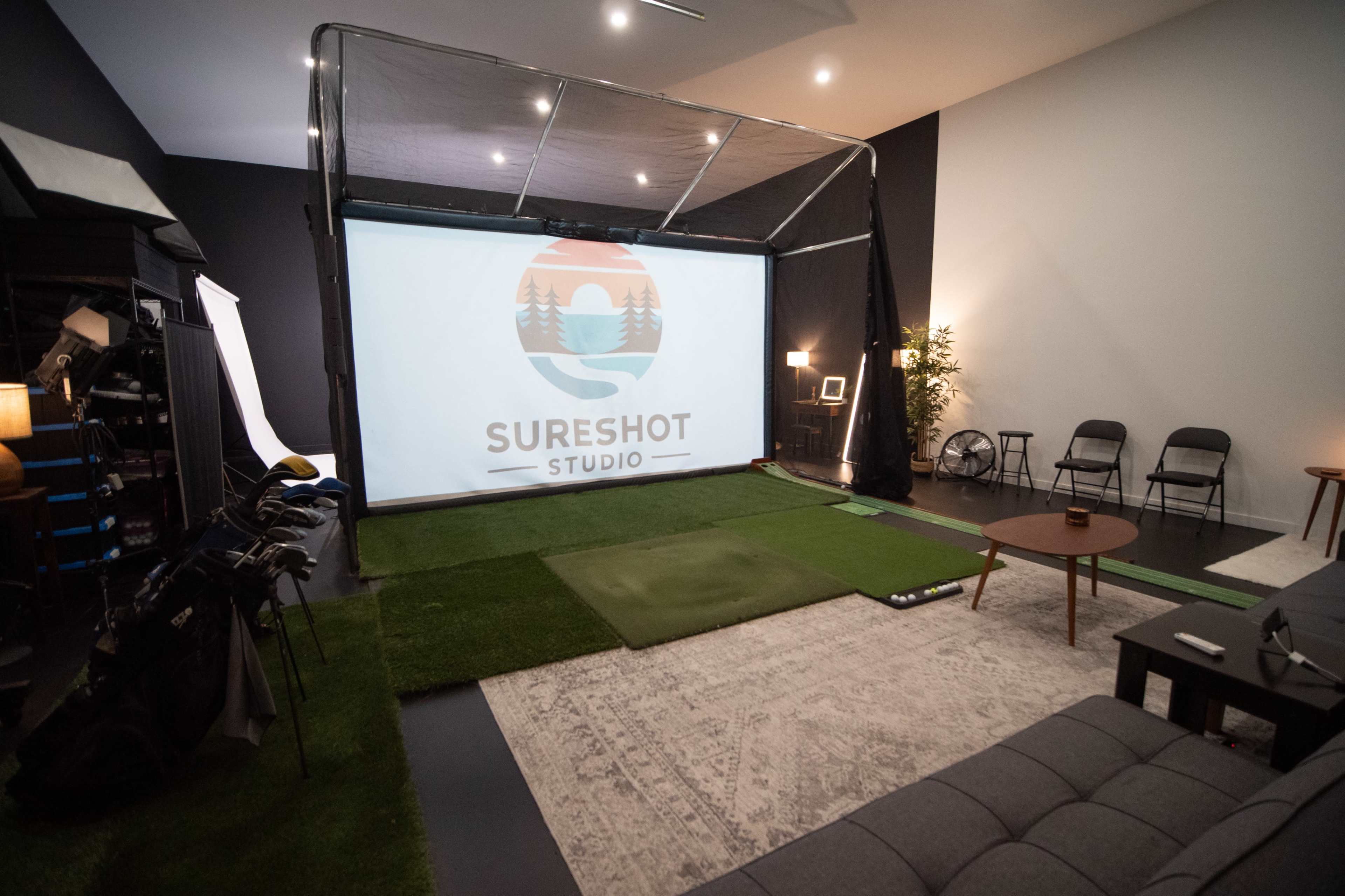 The image shows an indoor golf simulation studio with a large projector screen displaying "SURESHOT STUDIO," surrounded by golf equipment and a putting green.