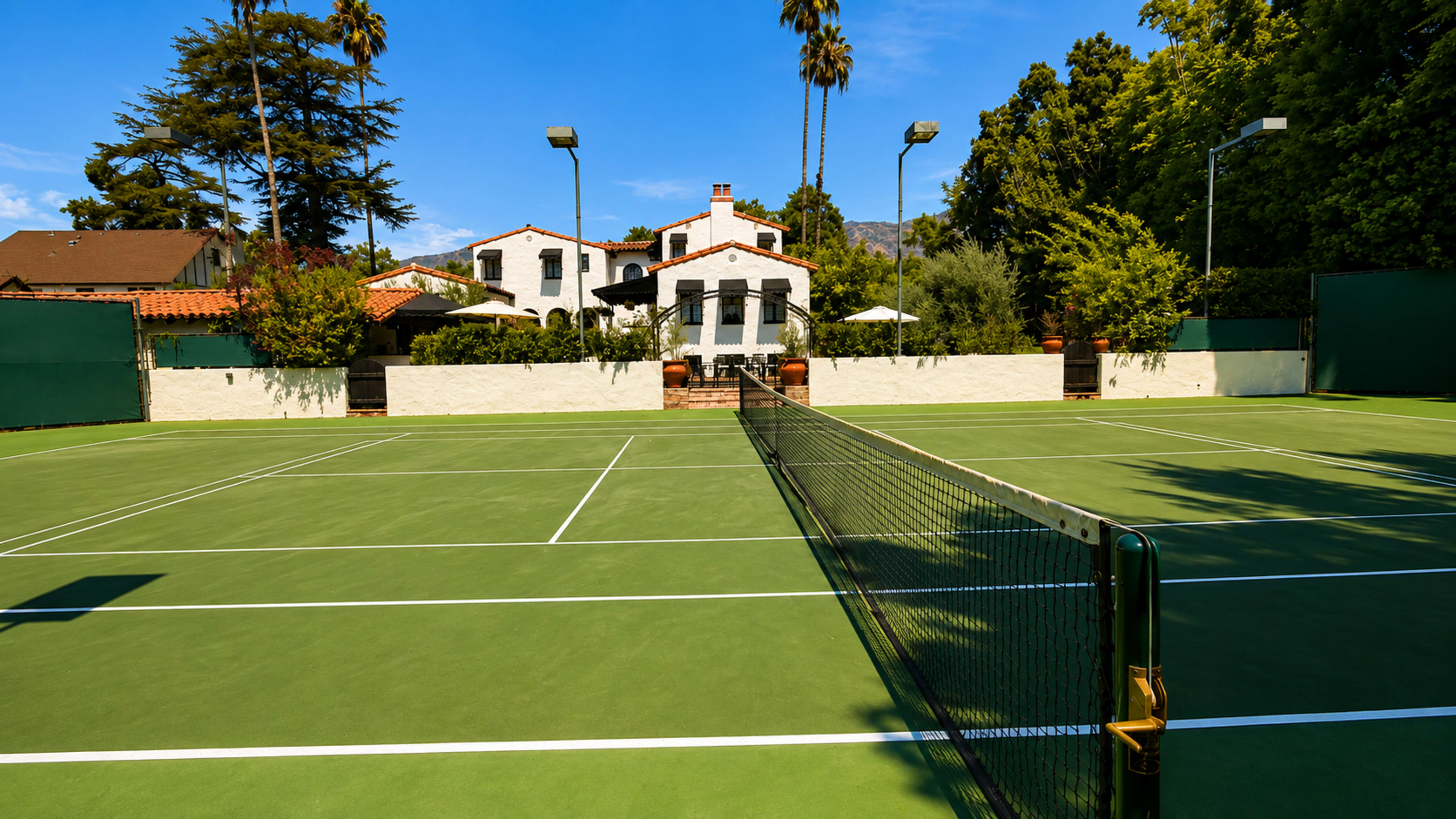 The image shows a tennis court with a net in the foreground, surrounded by trees and a building in the background.