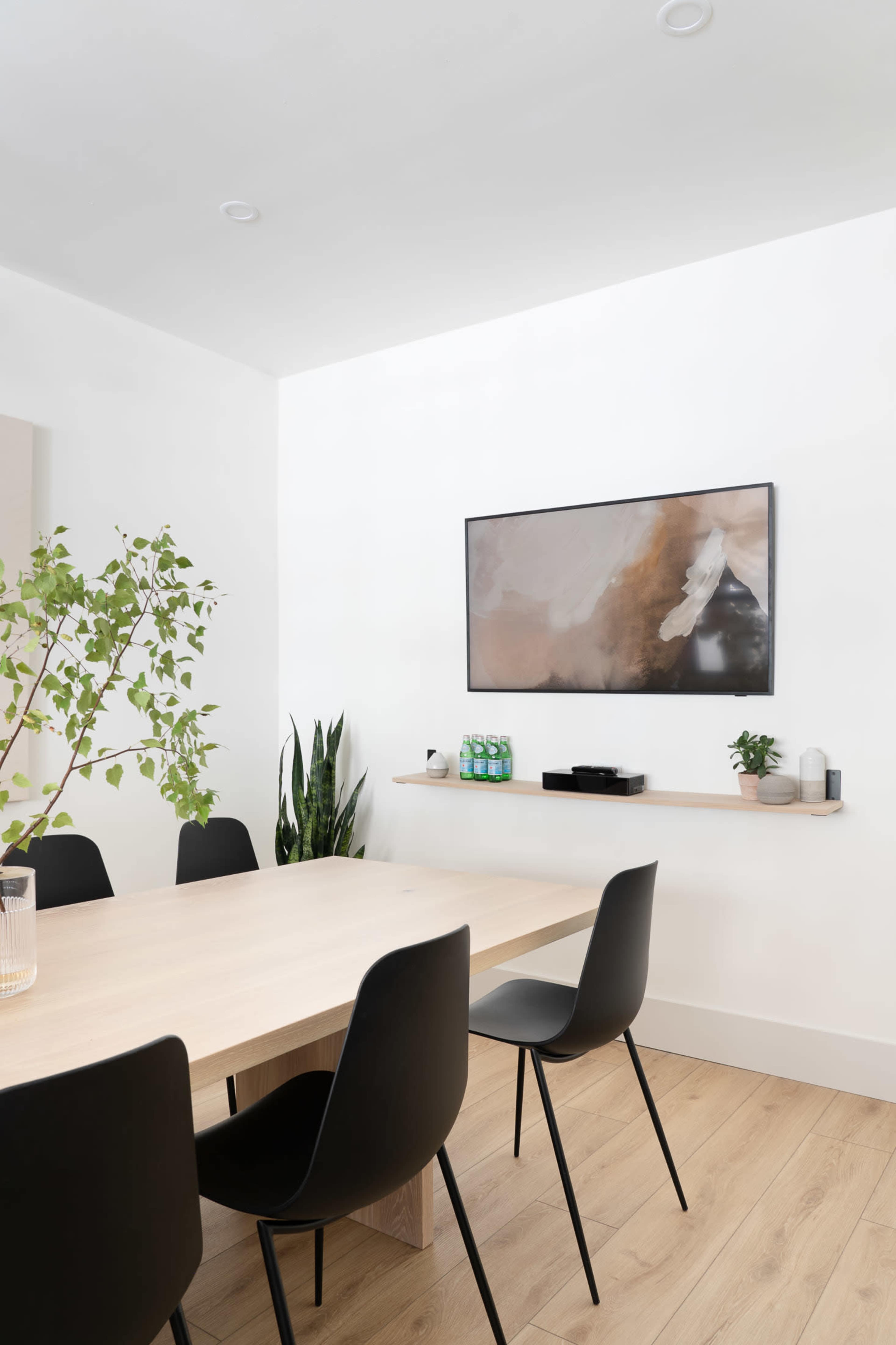 A modern dining area features a wooden table surrounded by black chairs, with a wall-mounted television and minimalist decor.