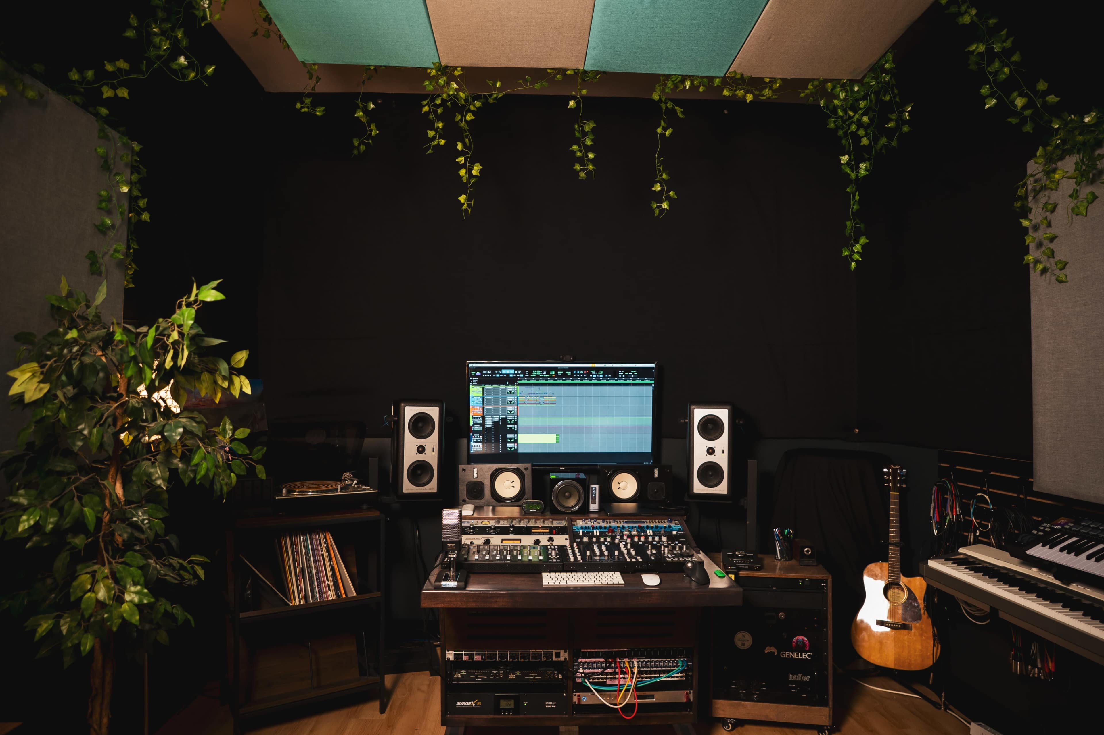 A music studio is equipped with a computer, audio equipment, speakers, an electric guitar, and decorative plants.