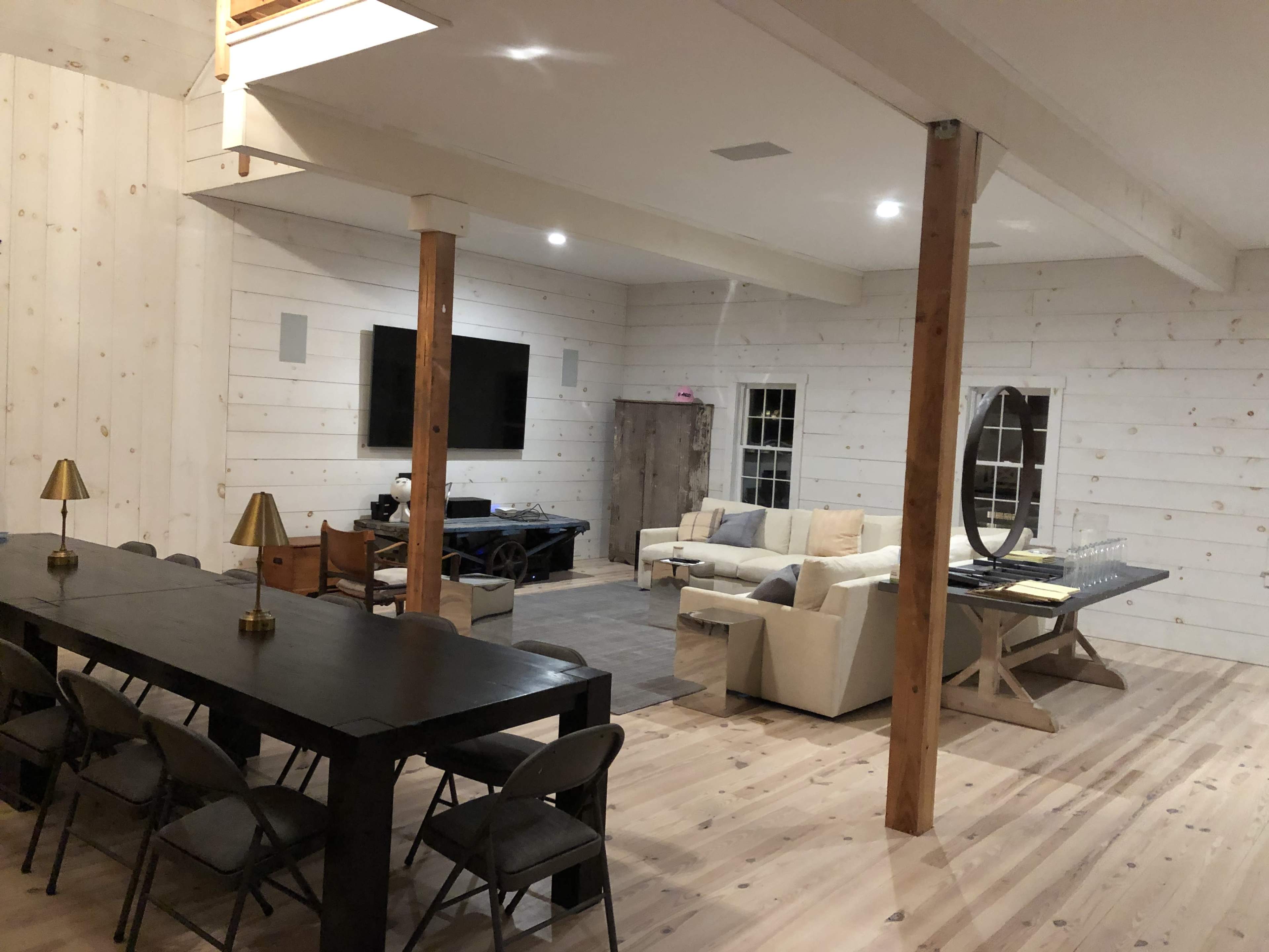 A spacious, modern interior with whitewashed wooden walls, a large dining table with chairs, a seating area with a light-colored couch, and a television mounted on the wall.