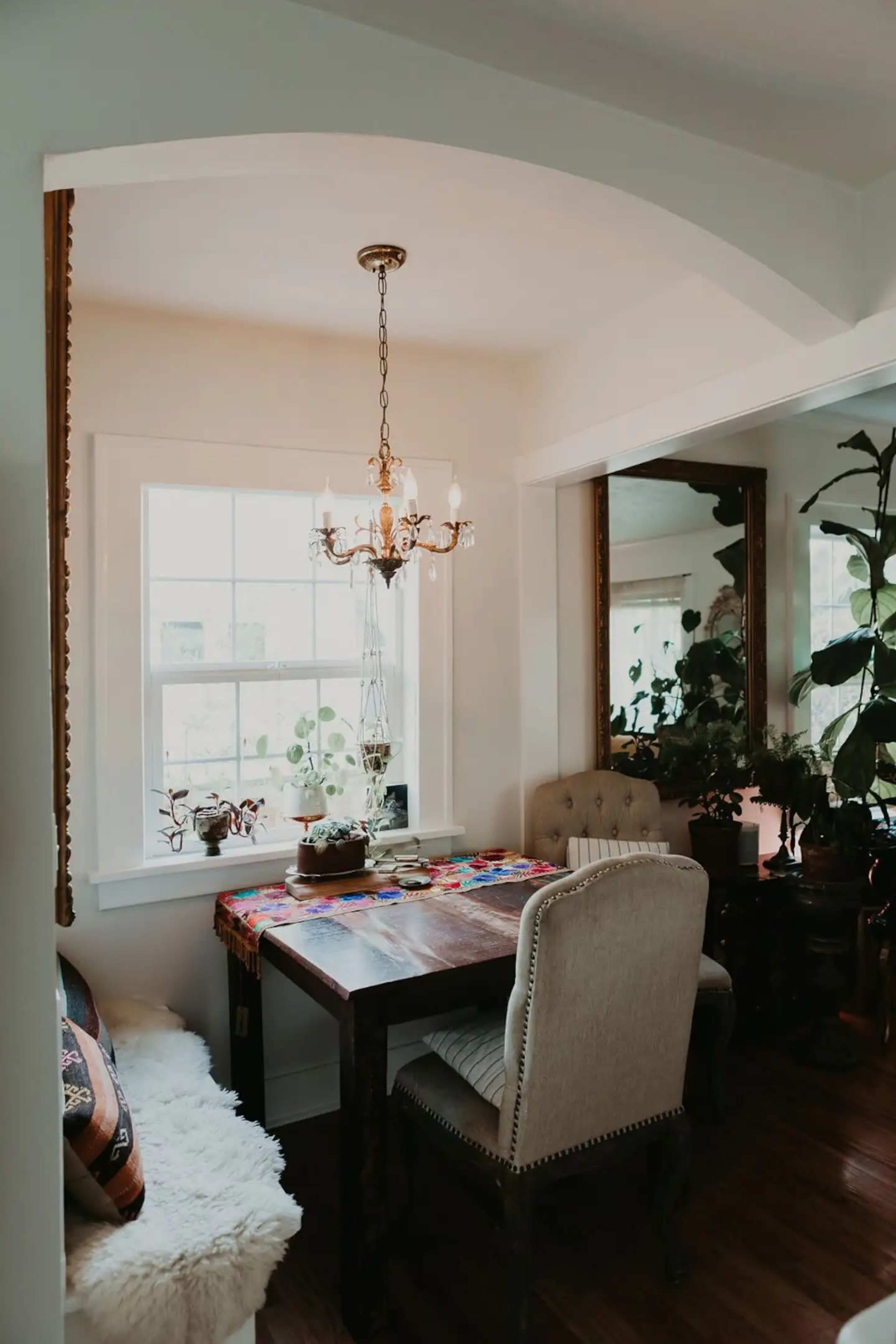 A small dining area features a wooden table with two chairs under a chandelier, next to large windows and surrounded by greenery.