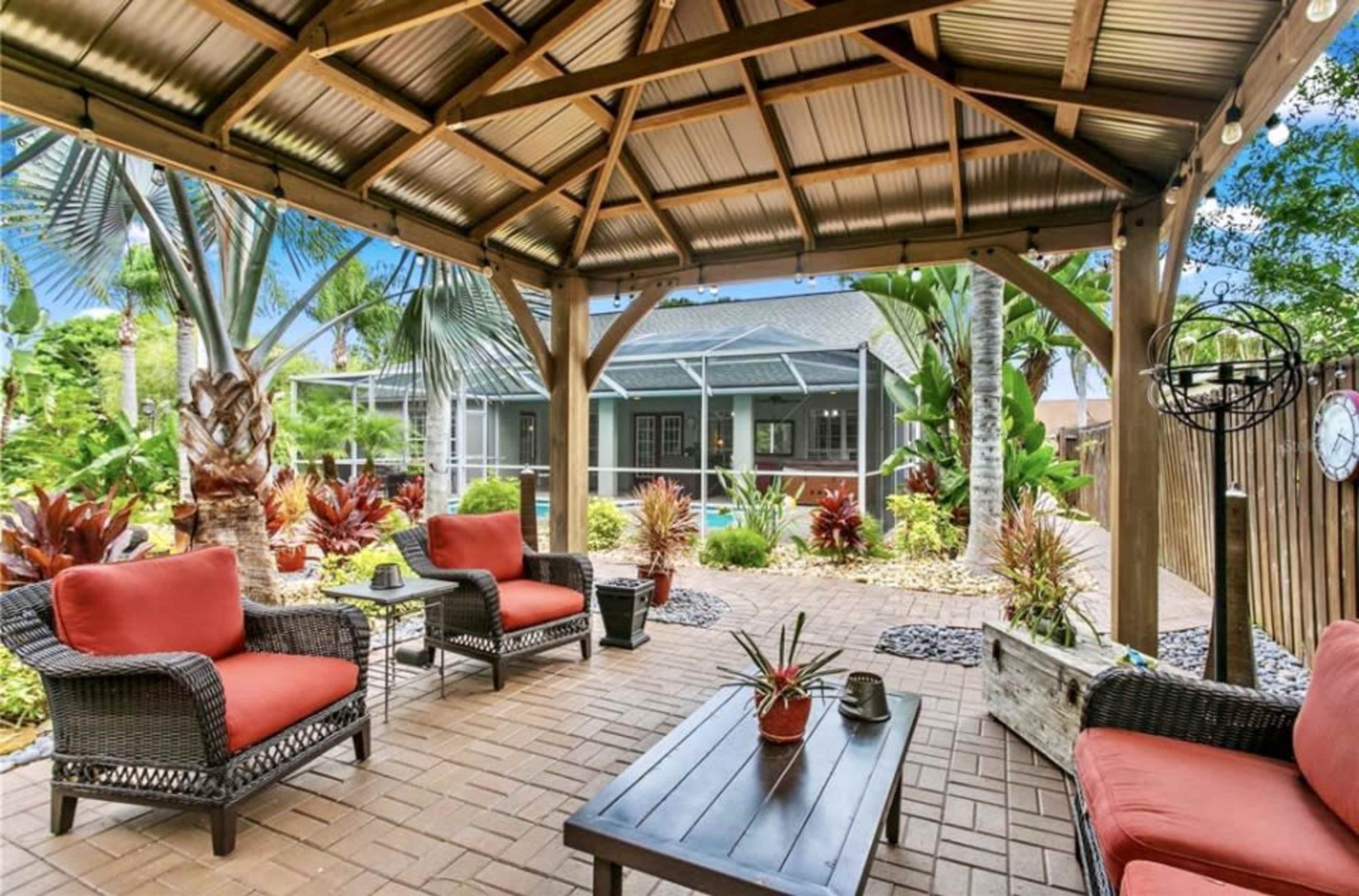 A covered patio area features wicker furniture with orange cushions, surrounded by tropical plants and landscaping.