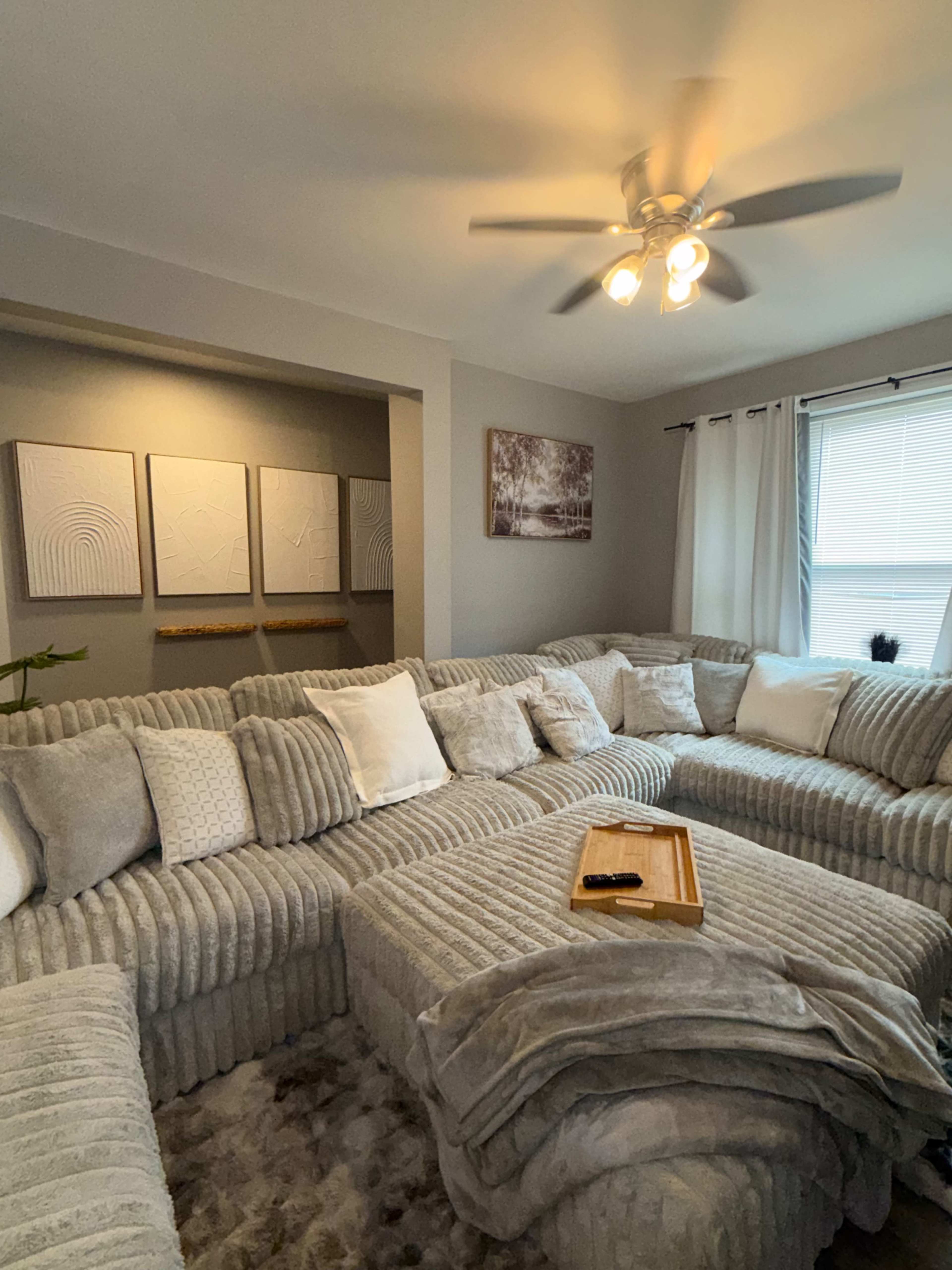 A large sectional sofa with gray fabric and multiple cushions is arranged in a cozy living room, accompanied by wooden decor and framed artwork on the walls.