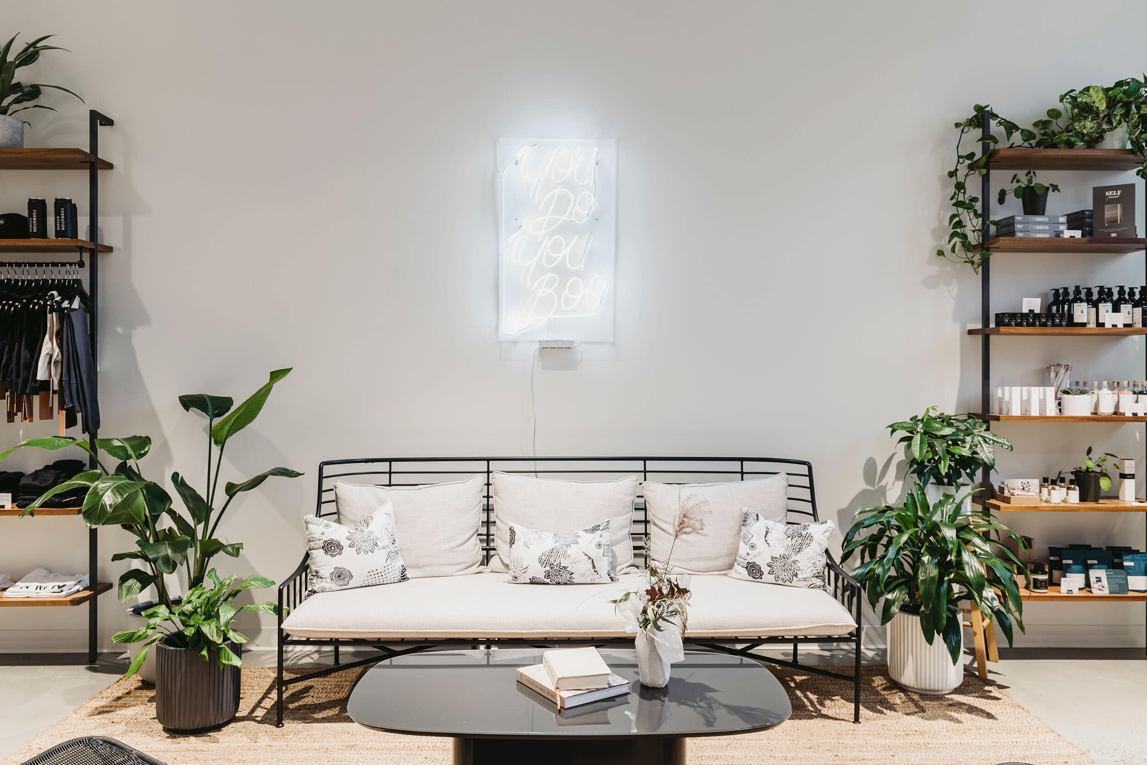 A minimalist indoor space featuring a black metal sofa with decorative pillows, a neon sign on the wall, and various potted plants alongside shelves filled with products.