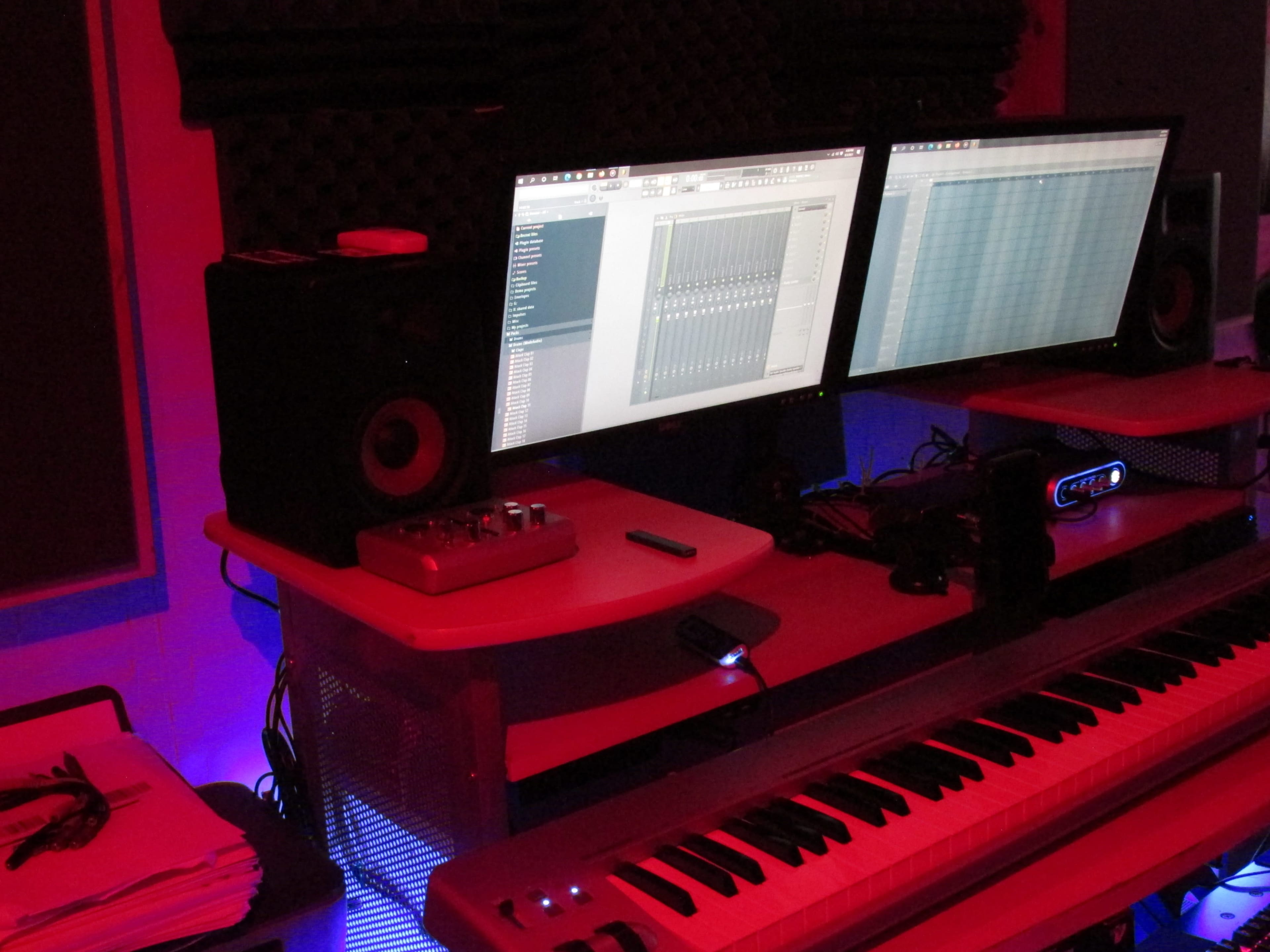 A music production setup features two monitors displaying digital audio software, a keyboard, and speakers in a dimly lit room.