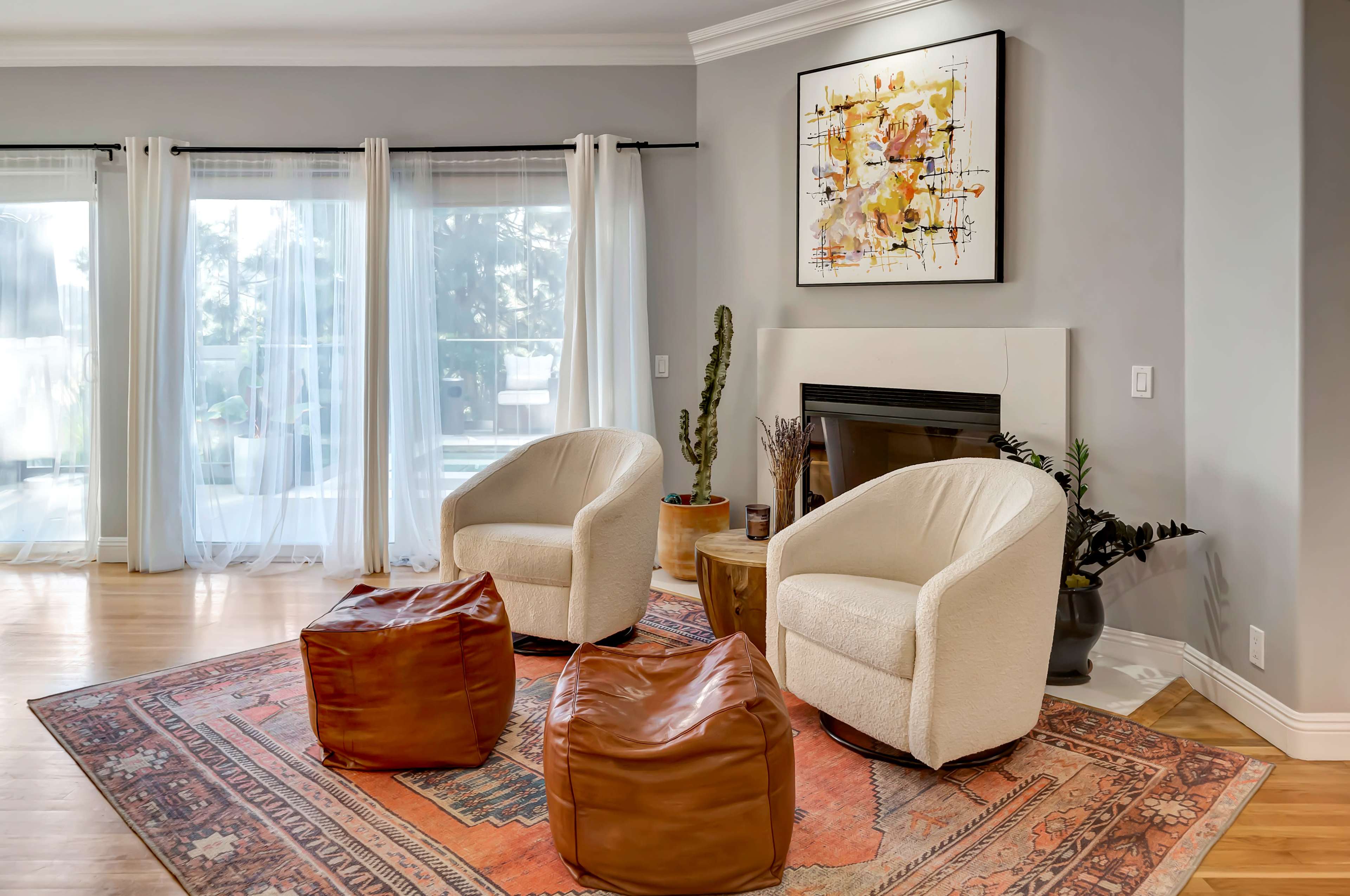 The image shows a spacious living room featuring two upholstered chairs, a small wooden table, and a decorative rug under a fireplace, with large windows draped in sheer curtains.