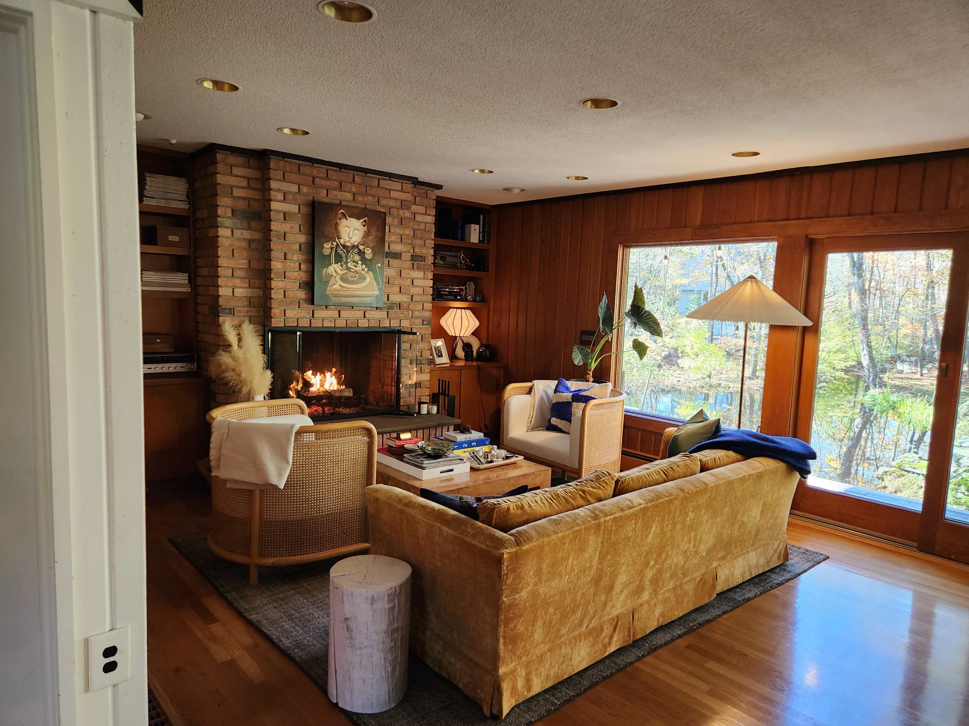 The image shows a cozy living room with a brick fireplace, a yellow sofa, wooden walls, a coffee table, and large windows overlooking trees.