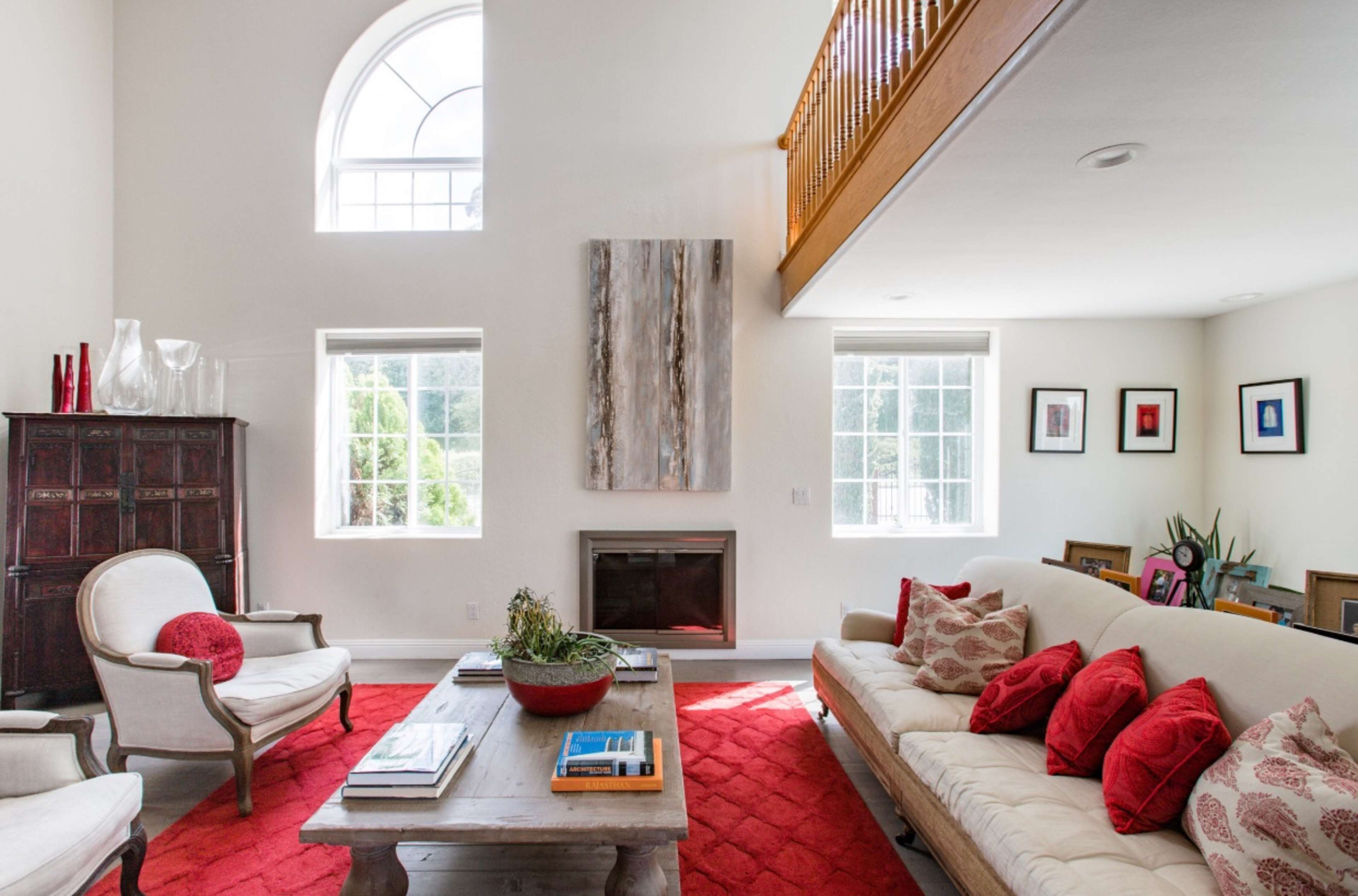 The living room features a large window above a beige sofa with red pillows, a wooden coffee table, and a red area rug, complemented by a balcony and several framed artworks on the walls.