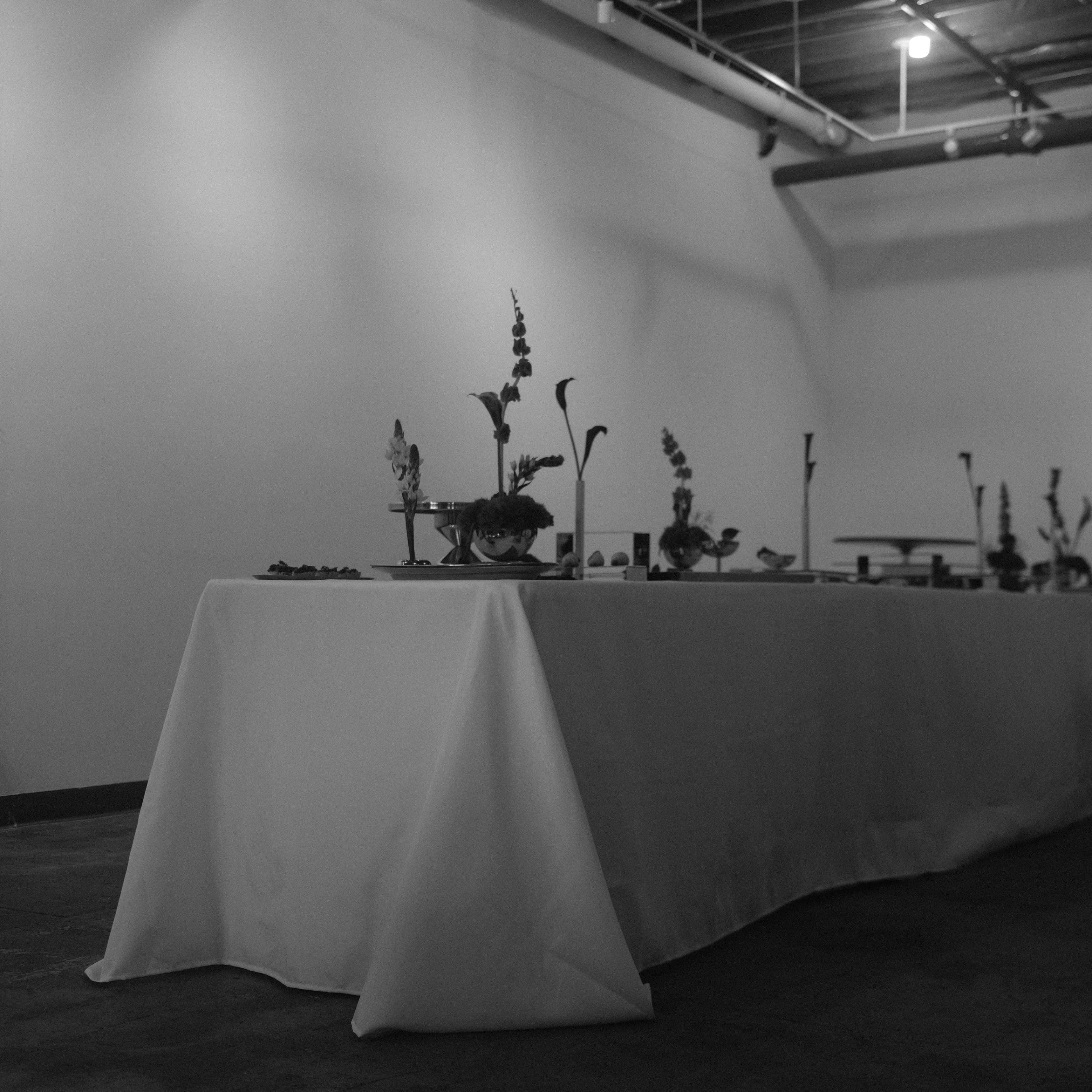 A long dining table, covered with a white tablecloth, is set with various decorative items and arrangements in a dimly lit room.