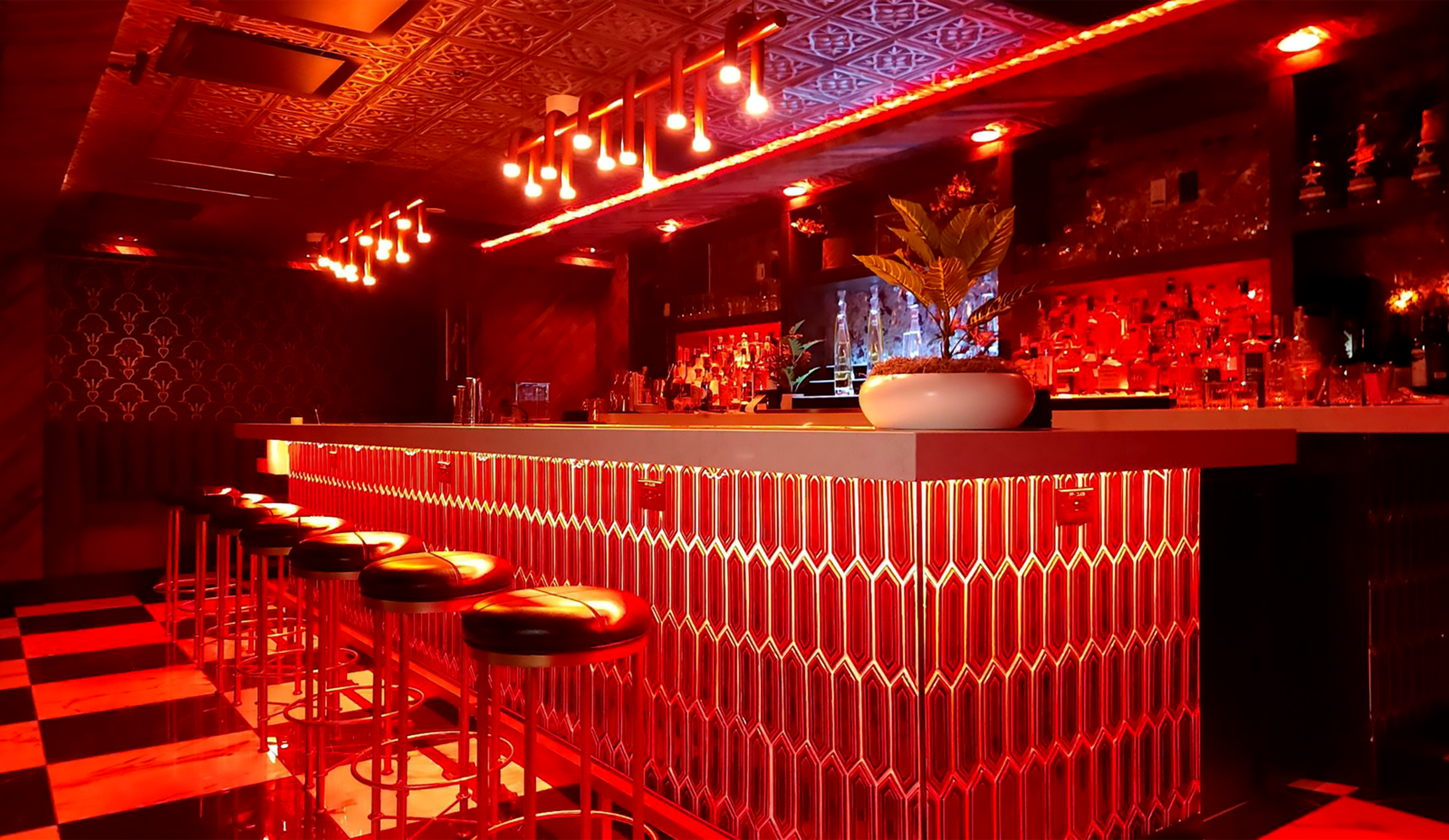 The image shows a dimly lit bar with a sleek red and black color scheme, featuring a long illuminated countertop and several high stools.