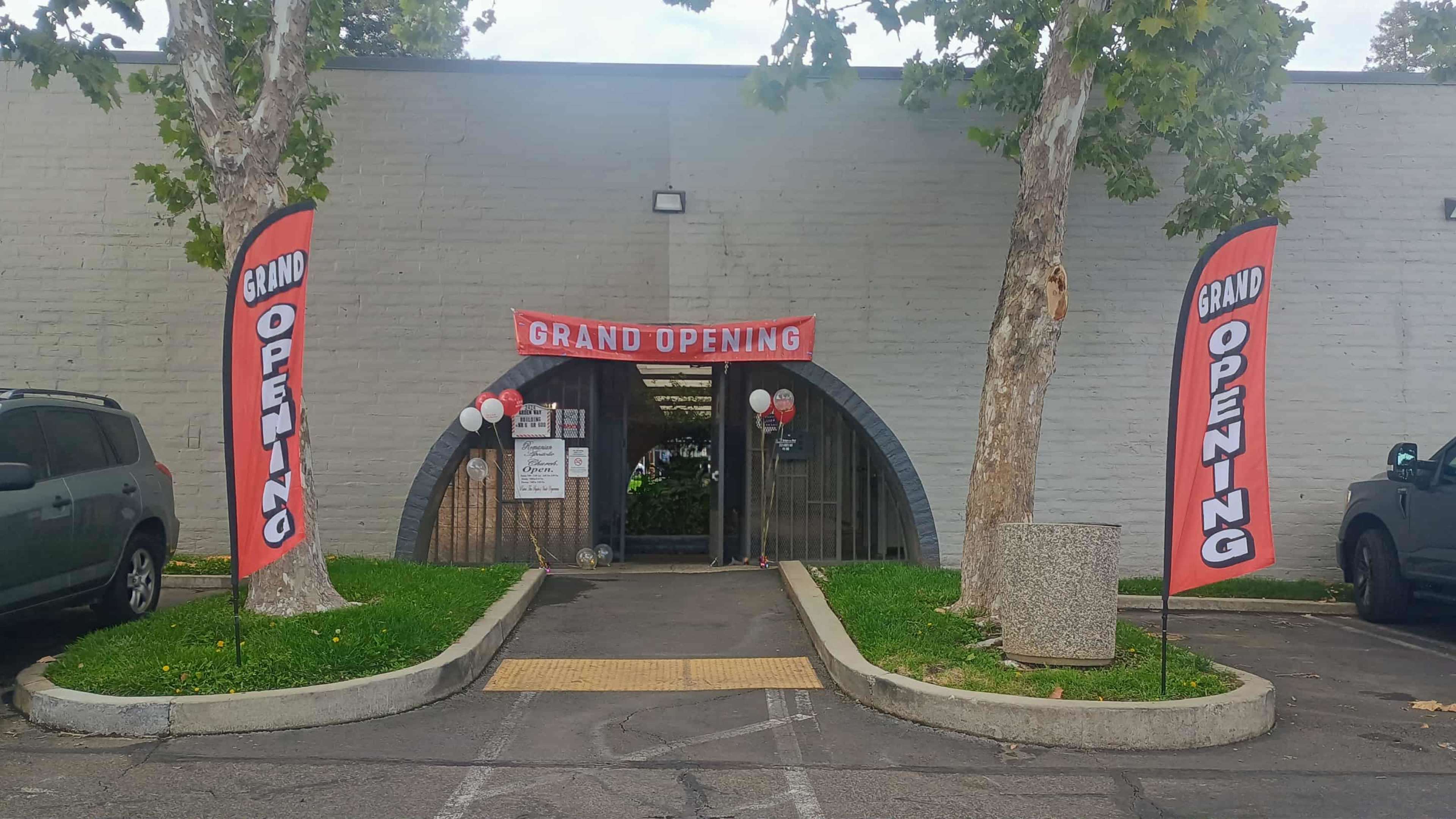 The image shows the entrance of a business with a "Grand Opening" banner and flags, flanked by two trees and parked cars.