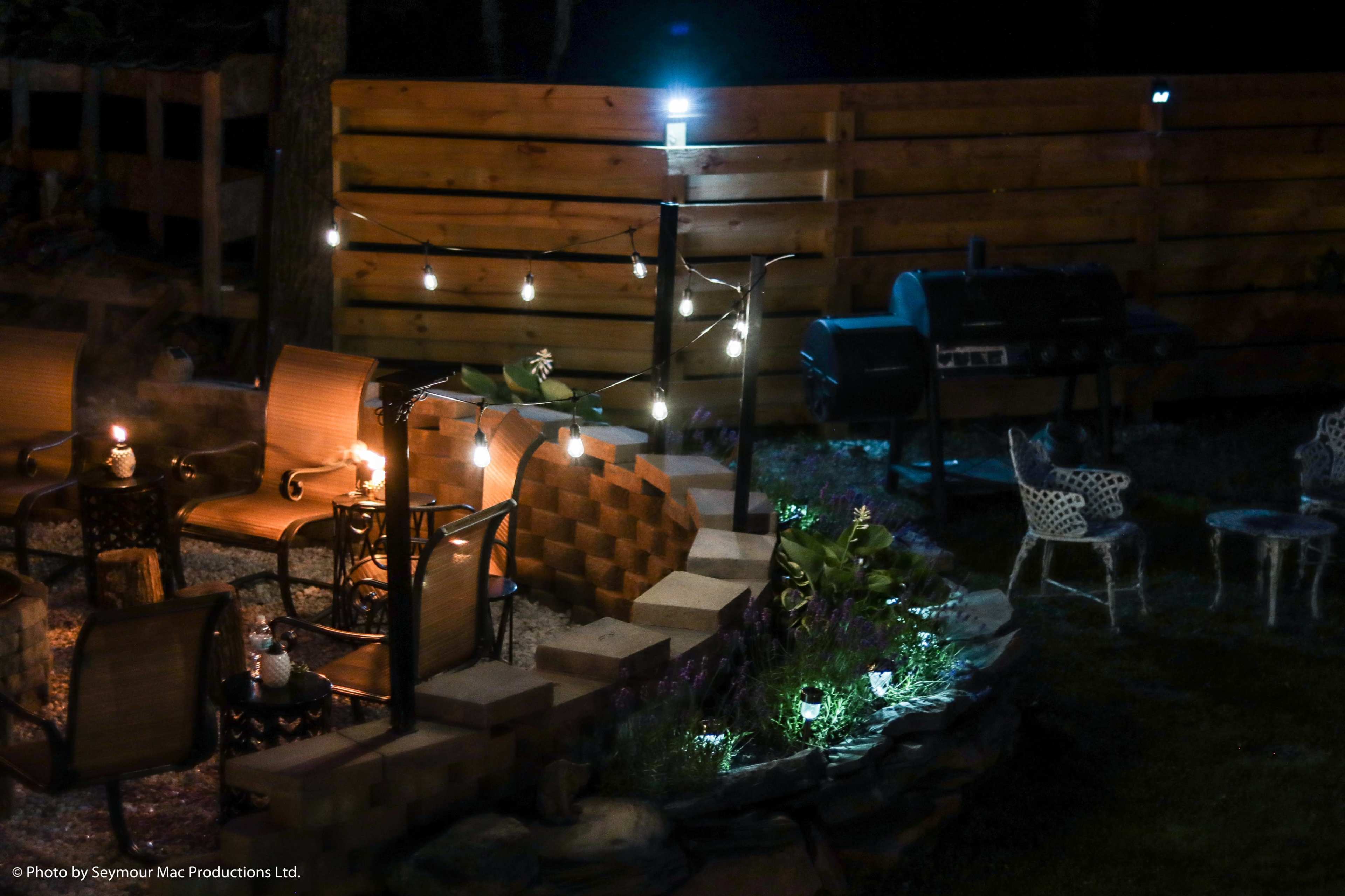 The image shows a well-lit outdoor patio area featuring a fire pit, seating arrangements, and a barbecue grill, surrounded by wooden fencing and decorative plants.