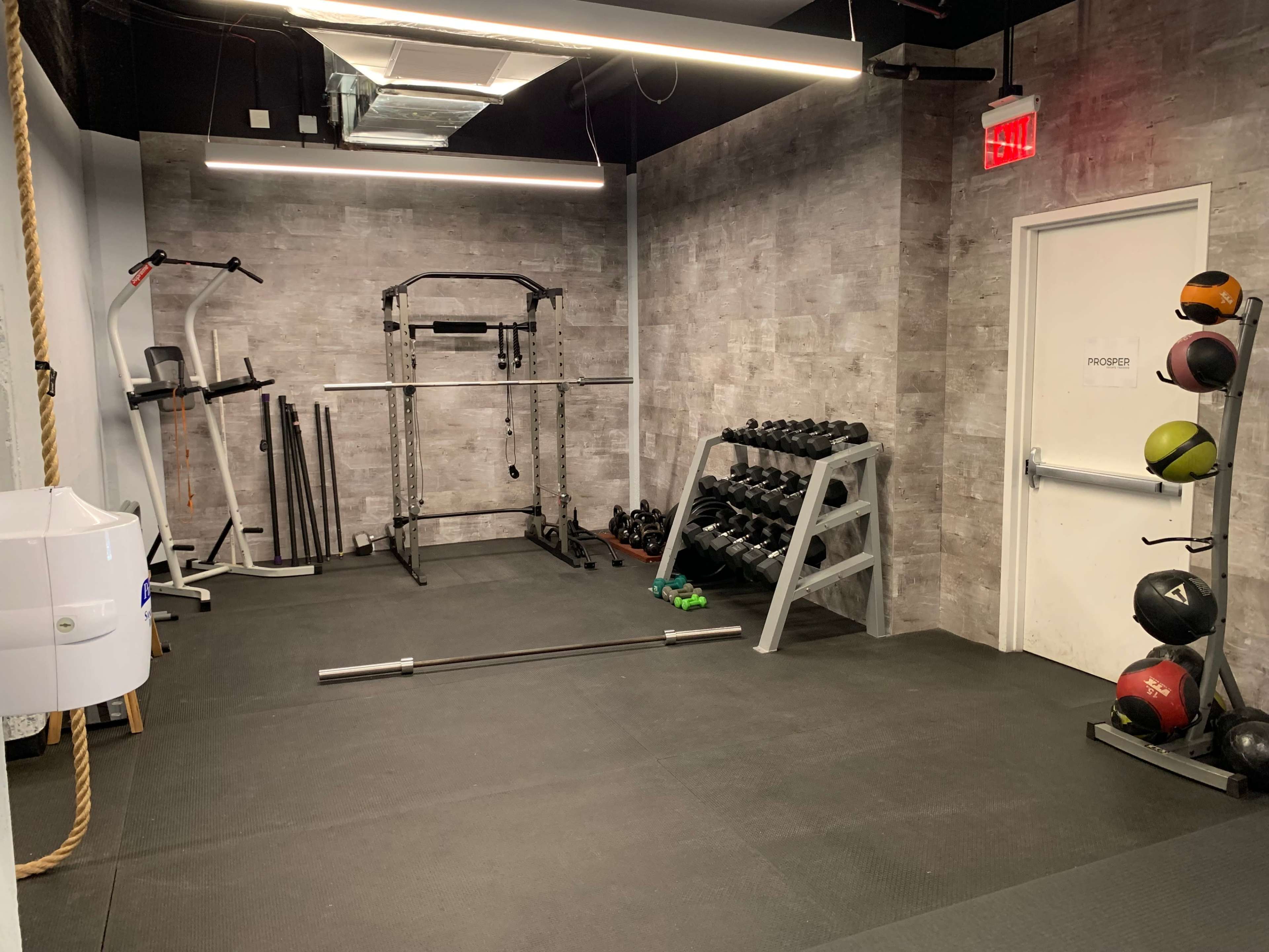 A small gym area featuring free weights, a squat rack, and exercise machines against a gray wall.