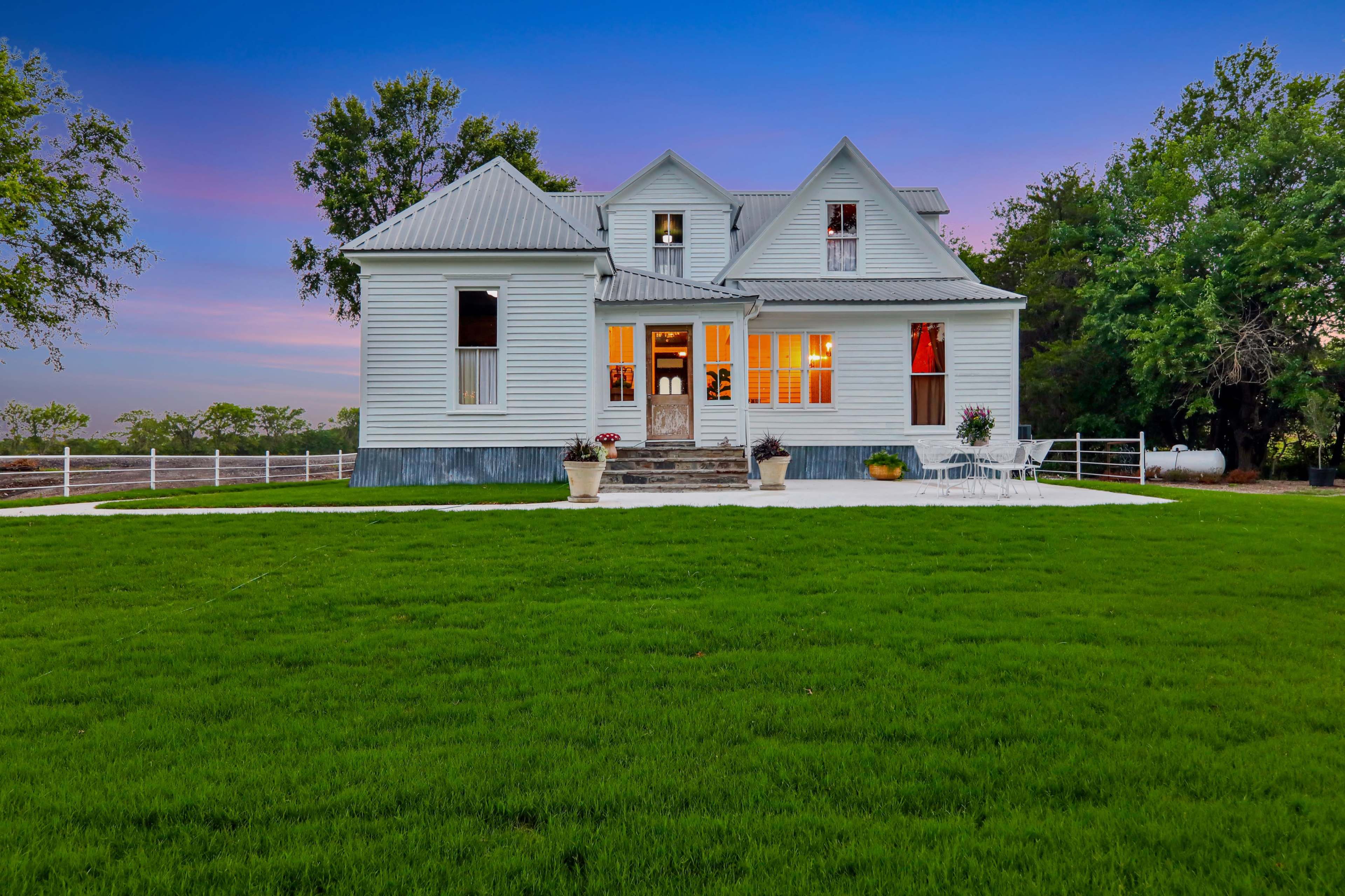 A two-story, white farmhouse with a metal roof features large windows and a spacious porch, set against a vibrant sunset and a lush green lawn.