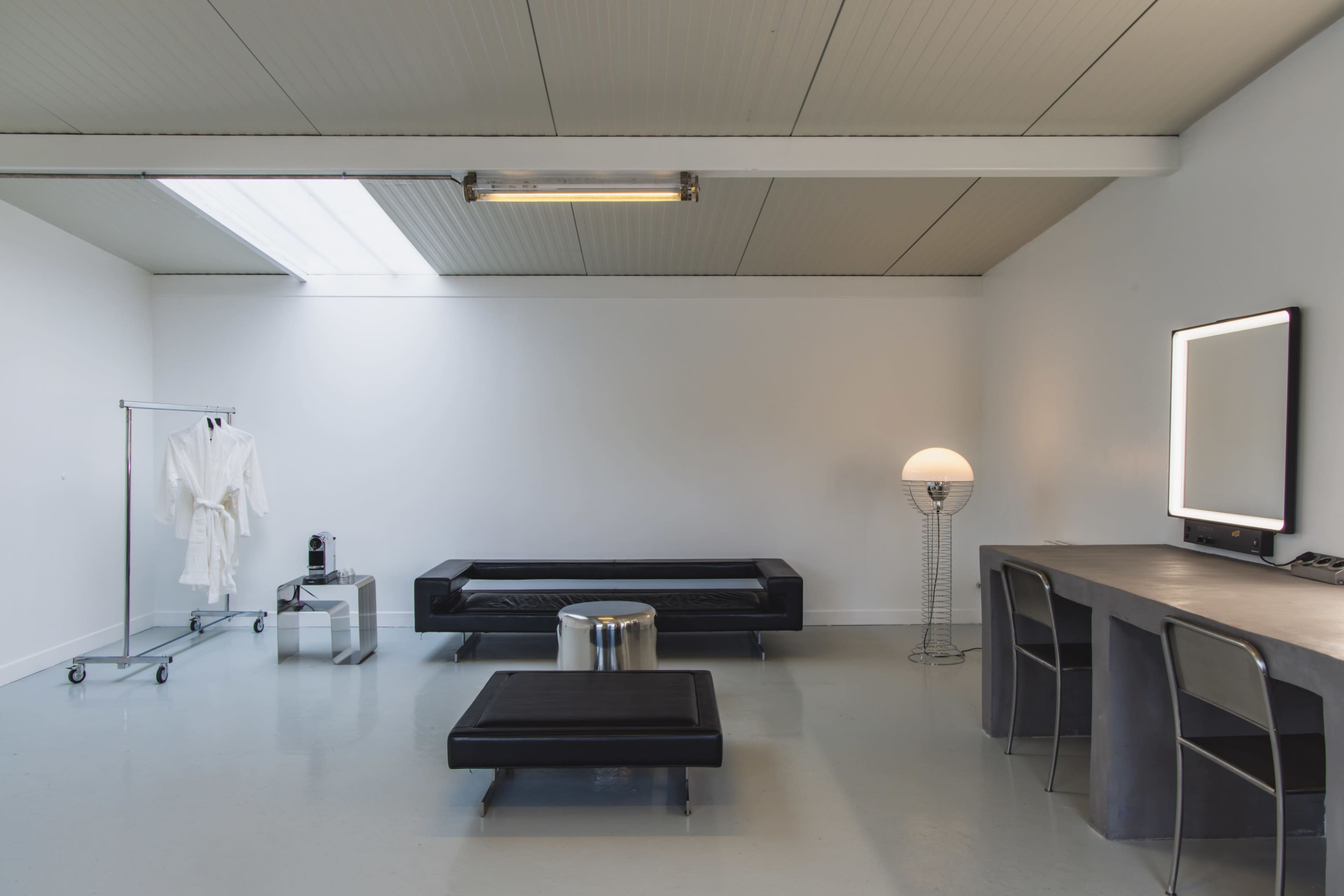 A modern, minimalist interior featuring a black sofa, a small coffee table, a clothing rack, and a large illuminated mirror with a vanity table.