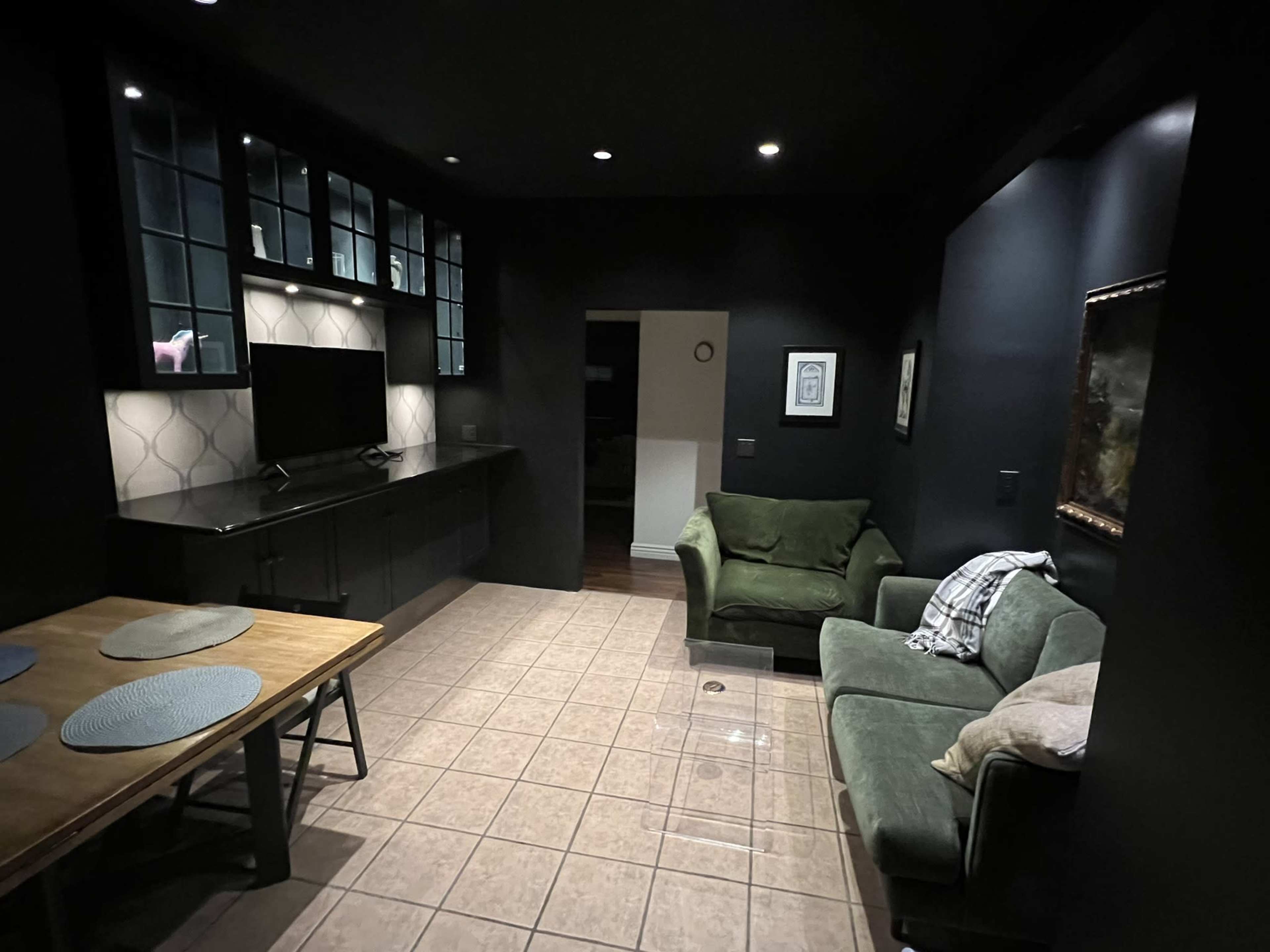 The image shows a dimly lit room with dark walls, a tiled floor, a green couch, a wooden table with placemats, and a TV mounted on a black cabinet.