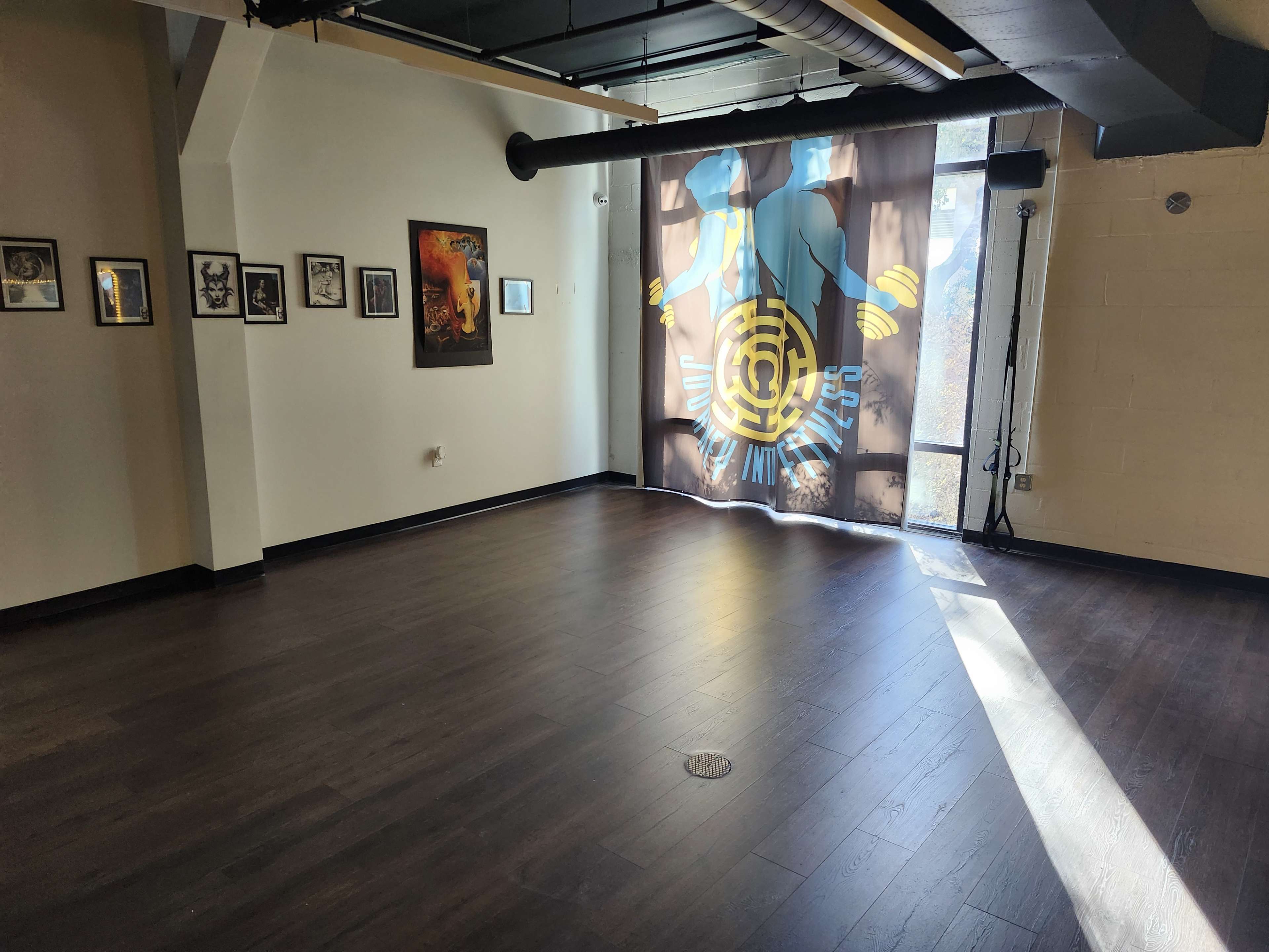 The image shows a spacious room with wooden flooring, adorned with framed artwork on one wall and featuring a large, decorative curtain with a design on the opposite wall.