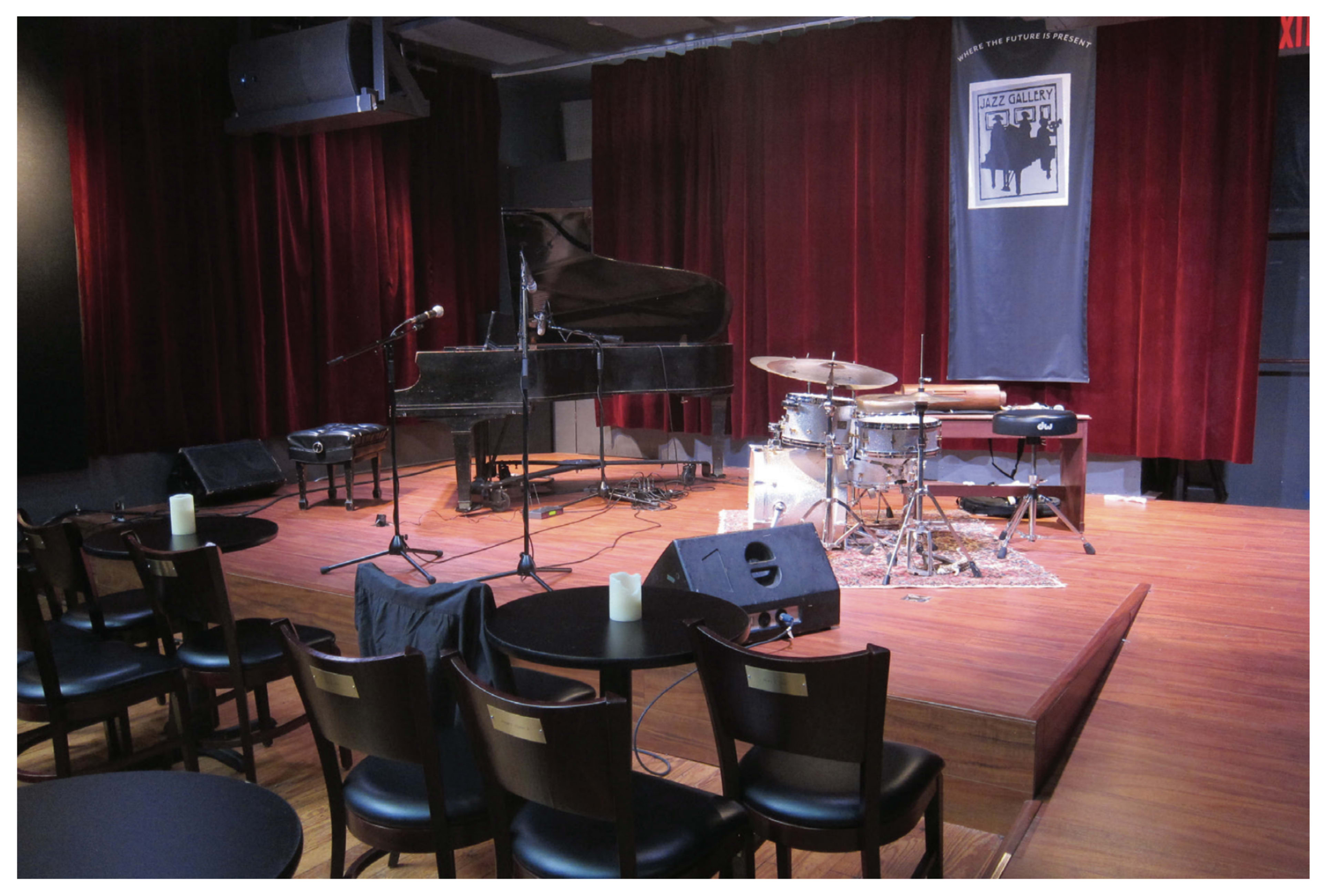 A small stage is set for a music performance, featuring a grand piano, a drum set, and microphones, surrounded by chairs and dim candlelight.