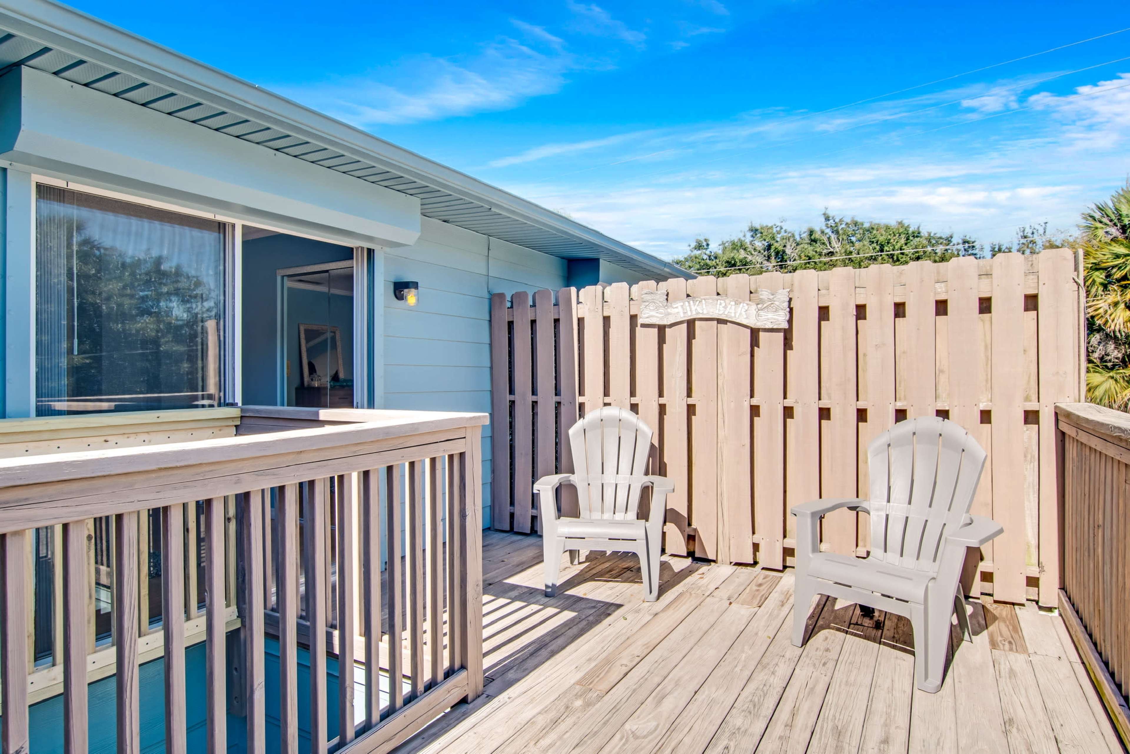 A wooden deck with two plastic chairs and a wooden fence enclosing the area, adjacent to a building with blue walls and a large window.
