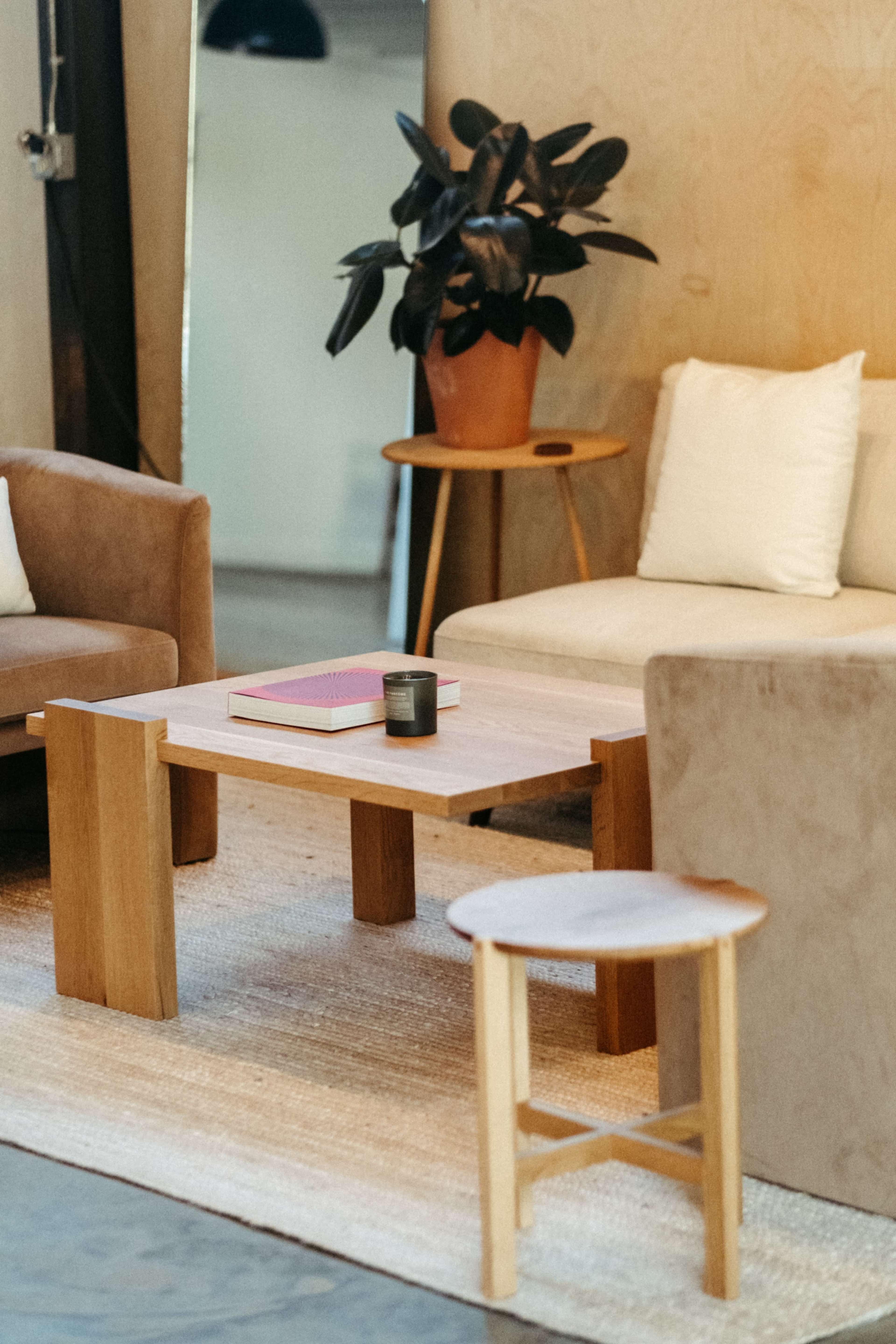 A minimalist living space features a wooden table with a book and a candle, surrounded by stylish sofas and a potted plant.