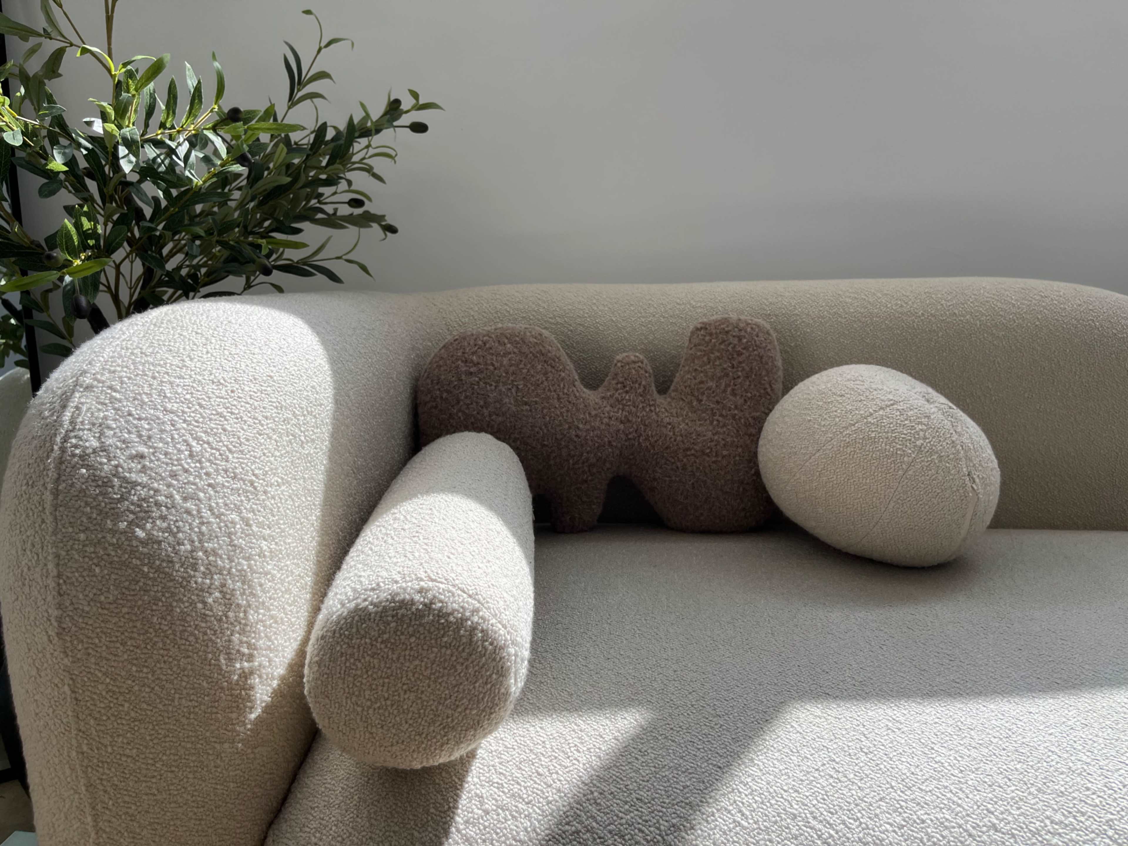 A beige sofa with cylindrical armrests features a decorative pillow shaped like the letter "W" and a round pillow.