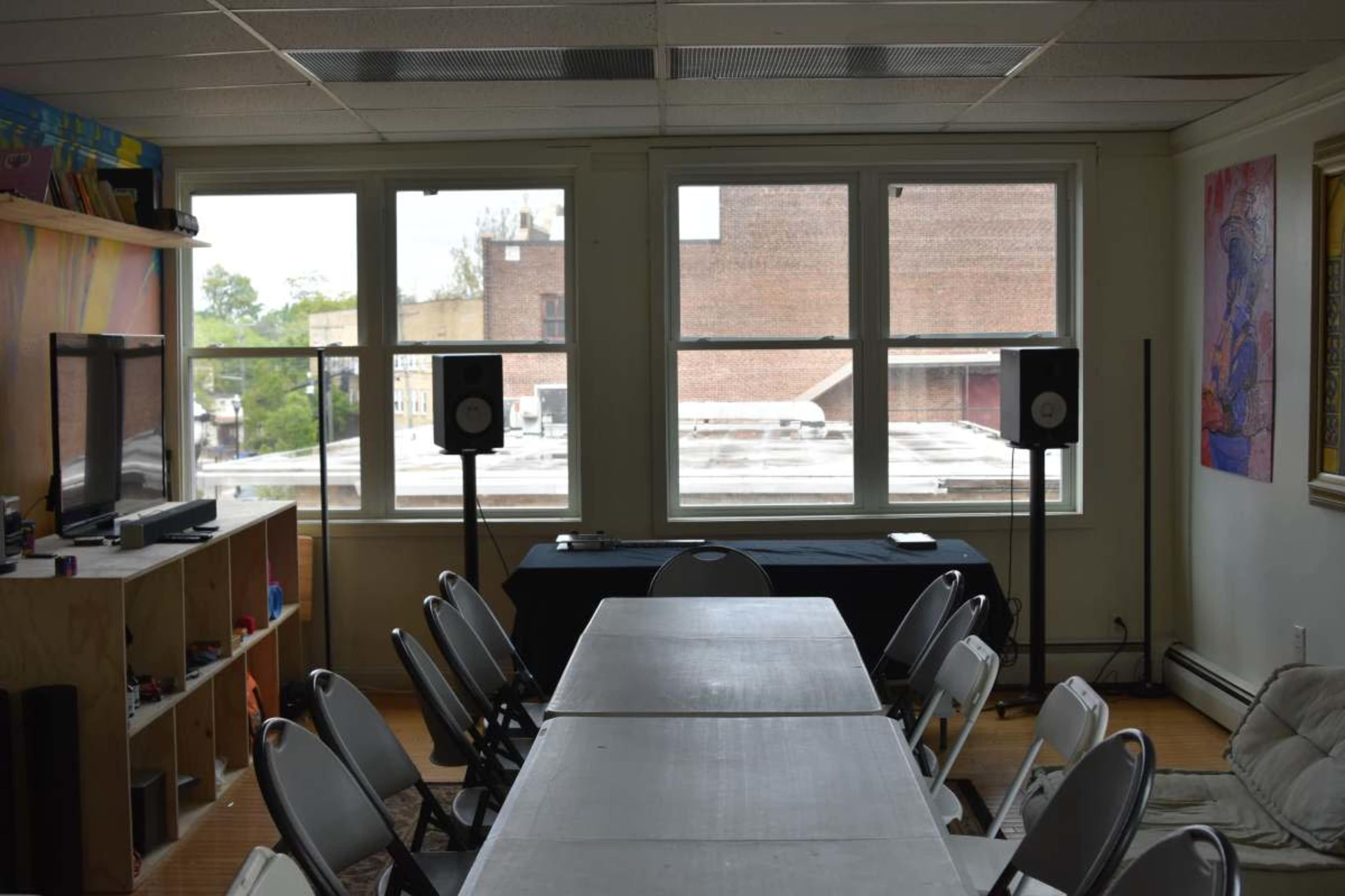The image shows a room with a long table surrounded by chairs, two speakers positioned near windows, and colorful artwork on the walls.