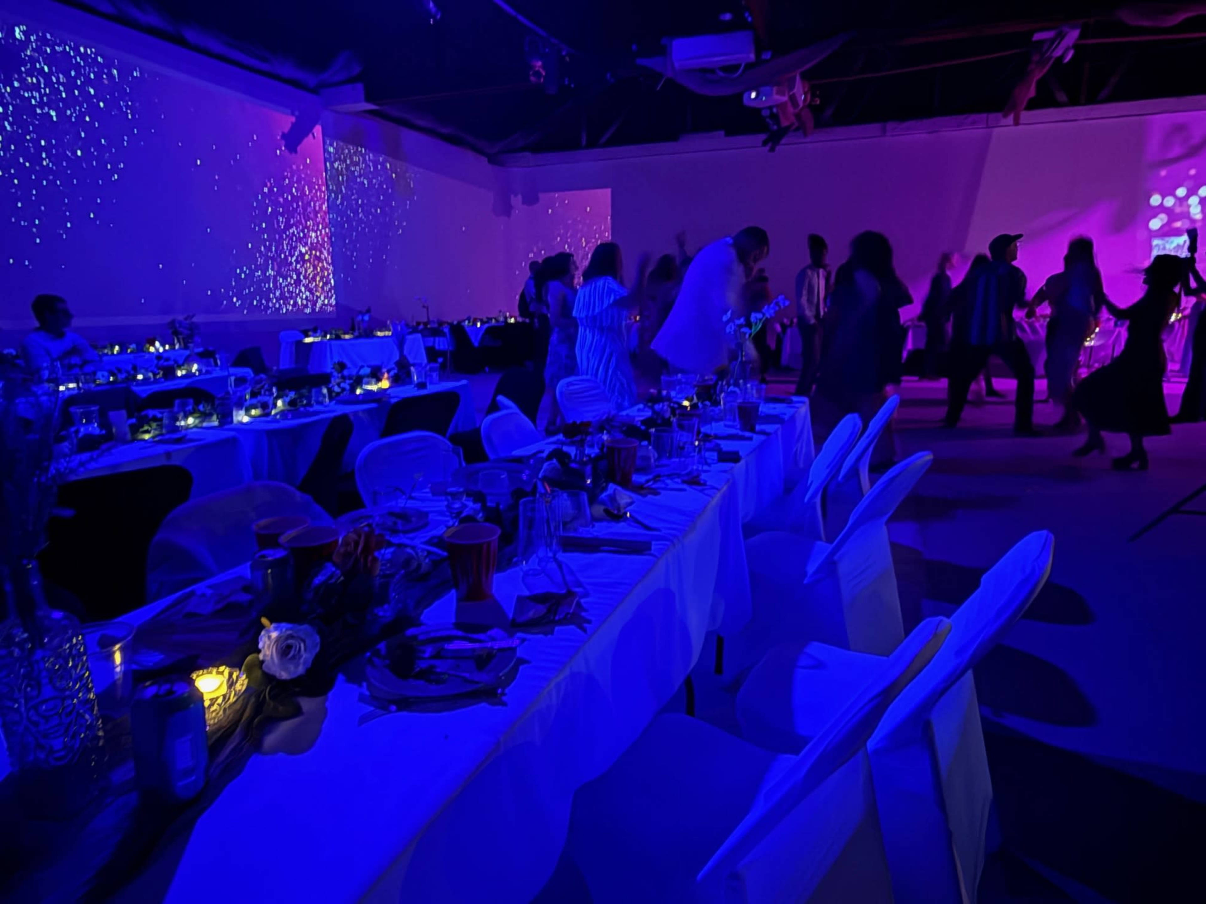 A banquet hall is set with tables covered in white cloths and illuminated by purple lighting, while guests are dancing in the background.