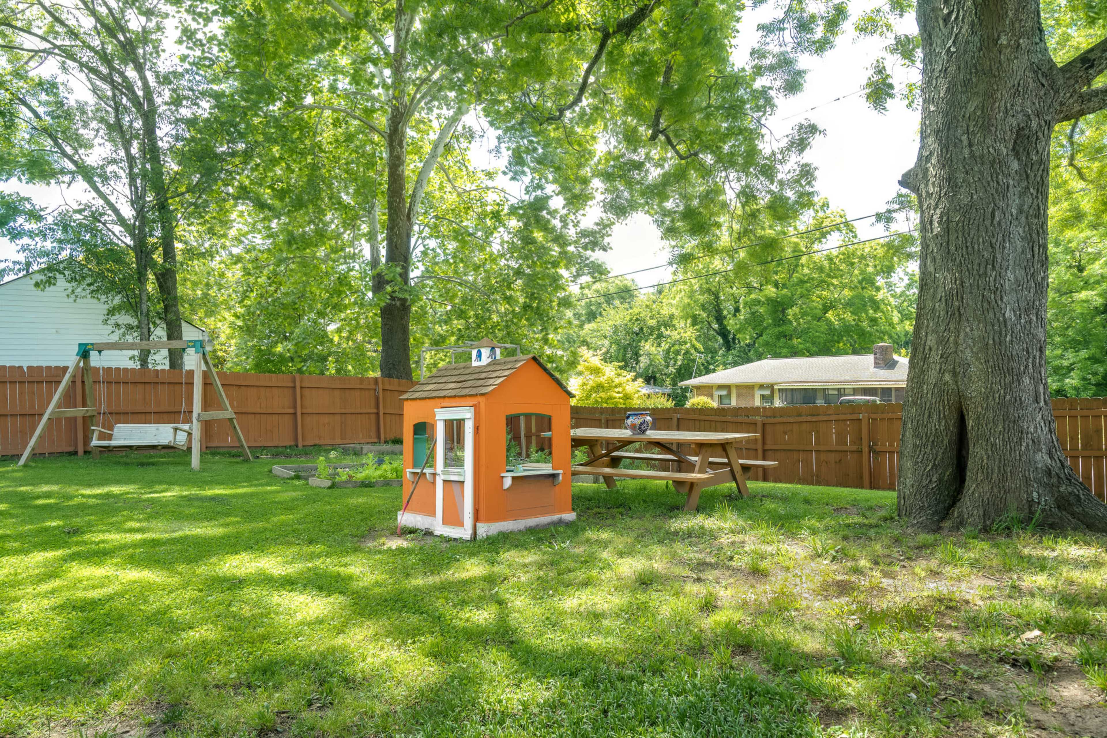 A backyard features a wooden playhouse, a picnic table, and a swing set surrounded by grass and trees.