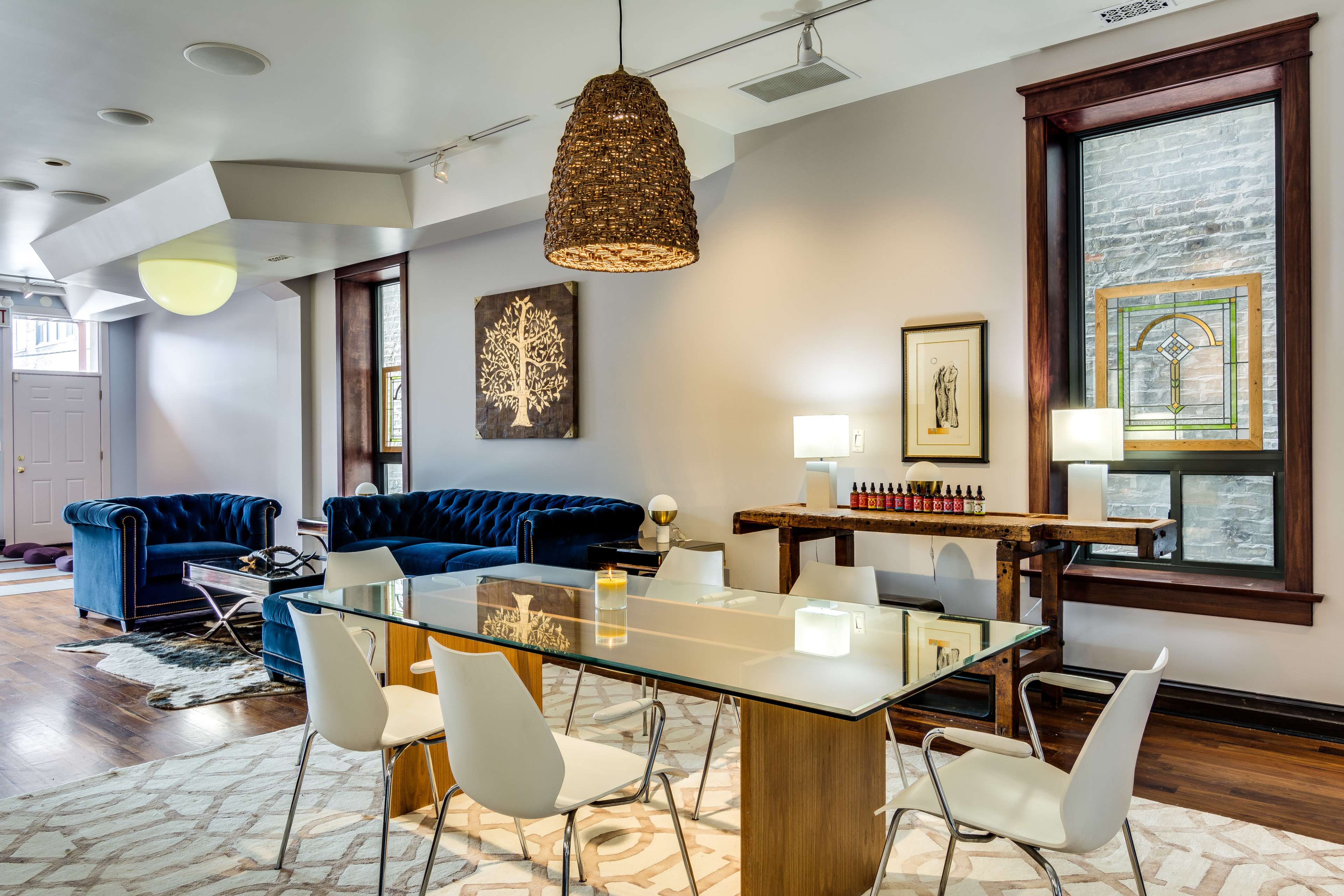 A modern interior space with a glass dining table surrounded by white chairs, a blue velvet couch, and decorative elements, including artworks and a woven pendant light.
