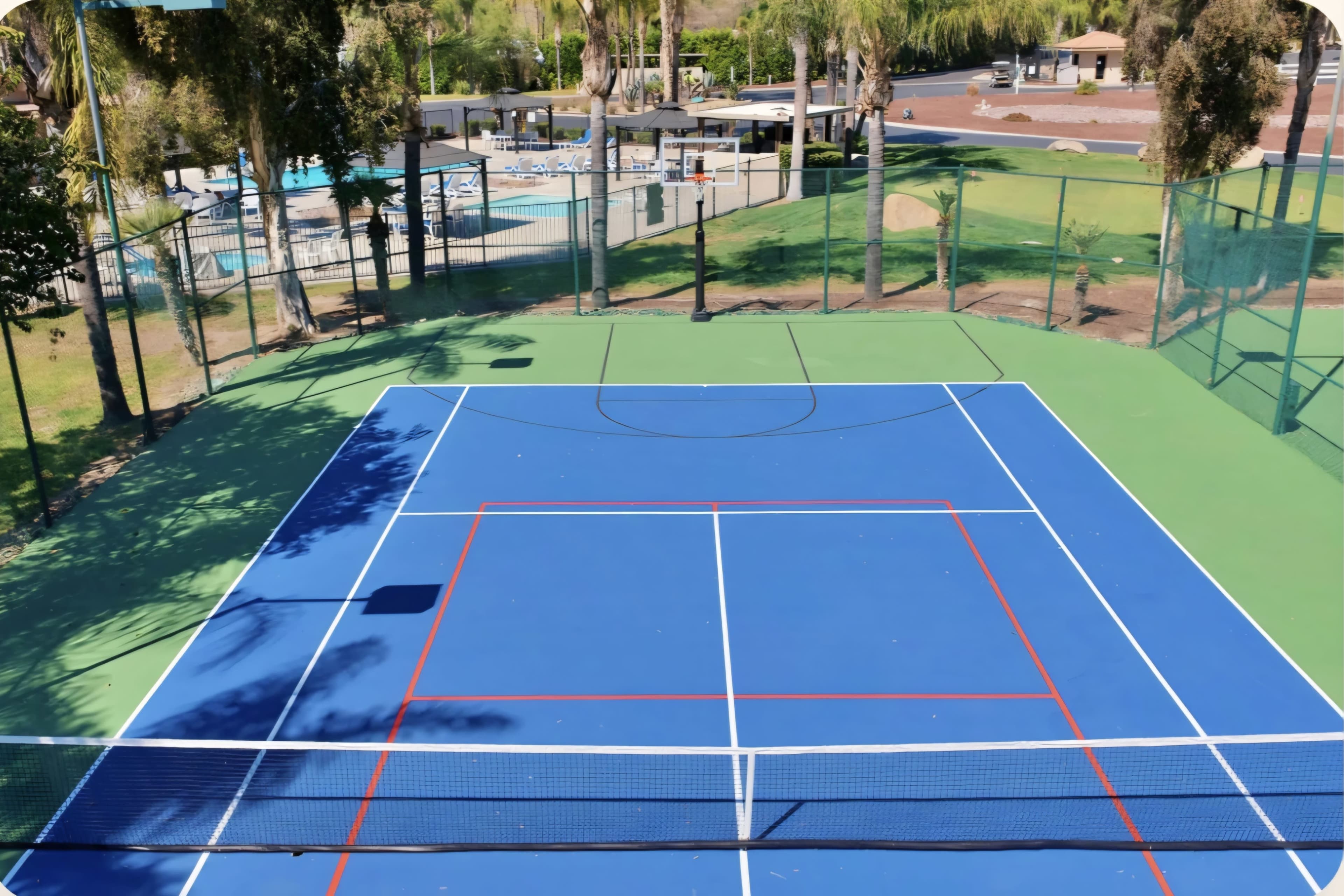 The image shows a well-maintained tennis court with a blue surface and red markings, surrounded by greenery and a pool area in the background.