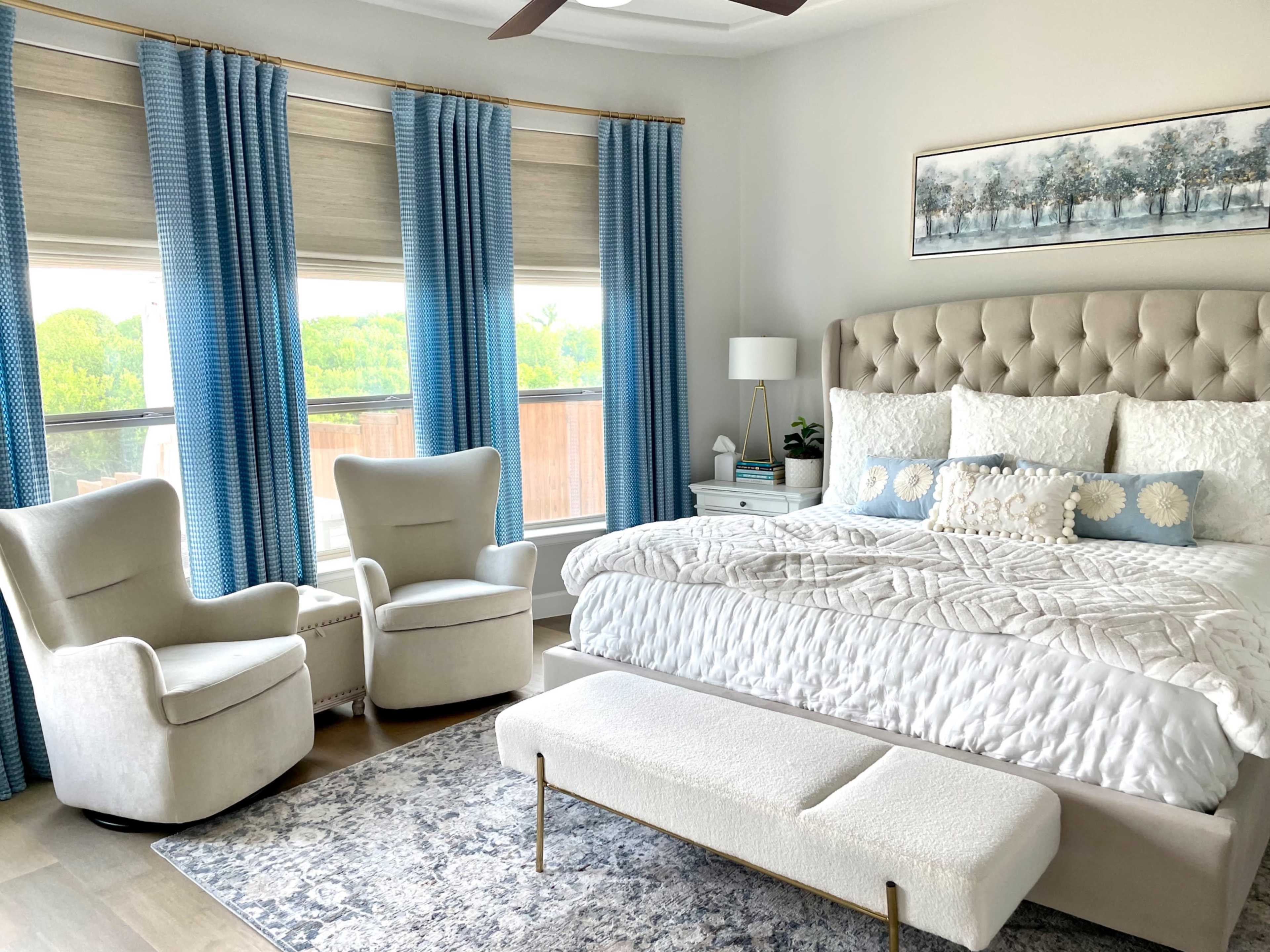 A well-furnished bedroom with a large bed, two armchairs, and large windows dressed with blue curtains.