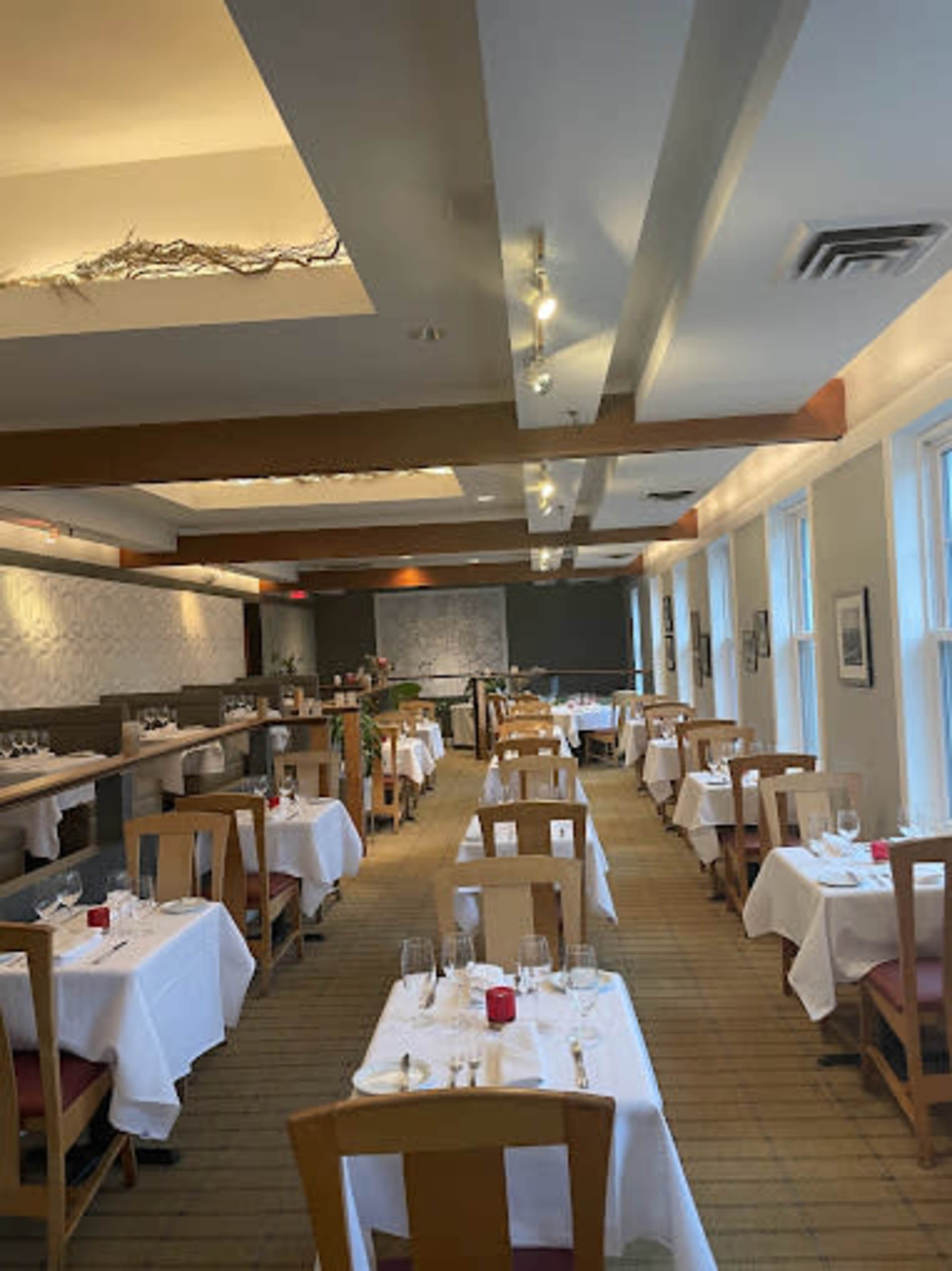 The image shows a formal dining room with white tablecloths on each table, arranged in rows with chairs, and soft lighting illuminating the space.