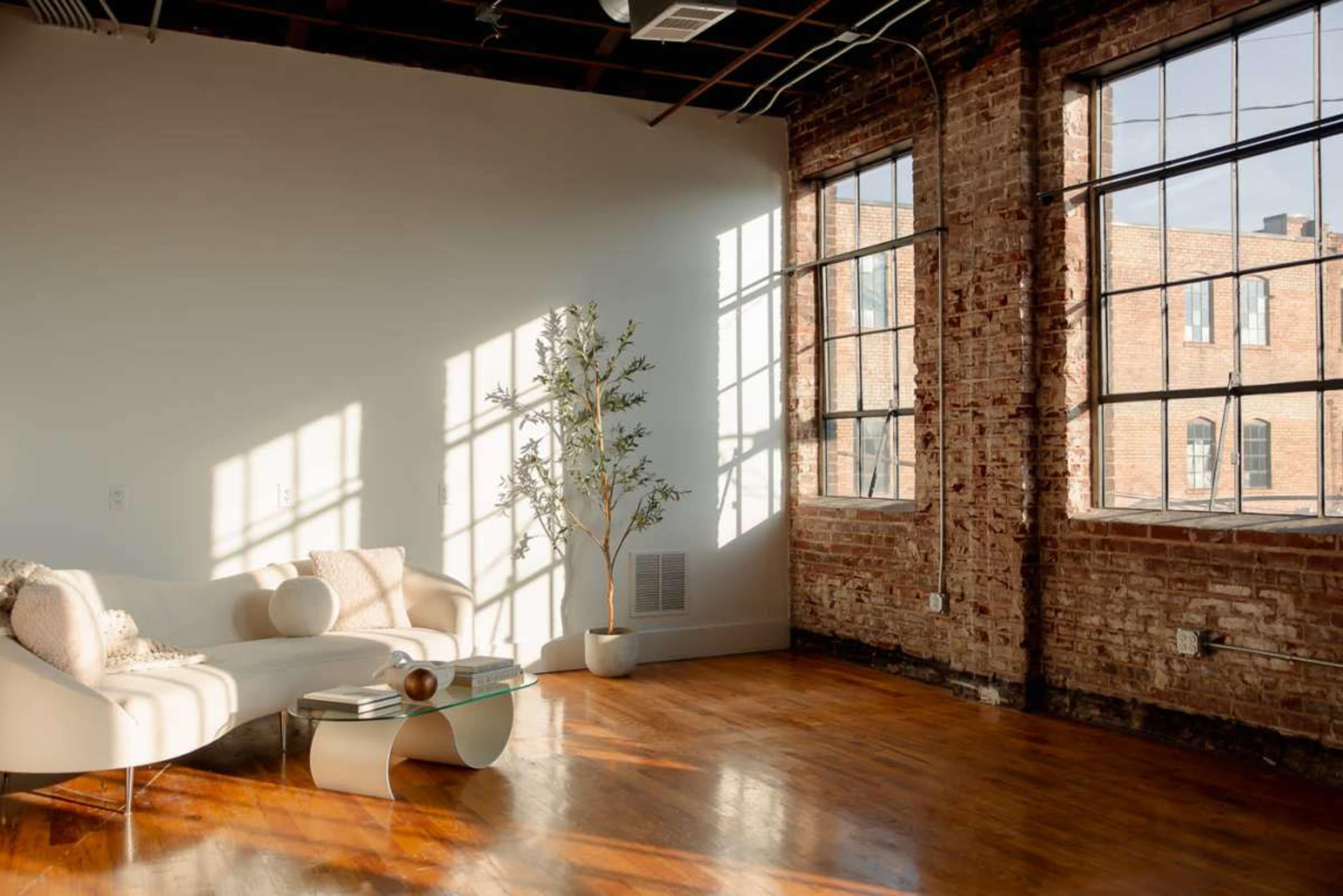 A bright, spacious room features large windows, exposed brick walls, a light-colored curved sofa, and a small table with a decorative plant.