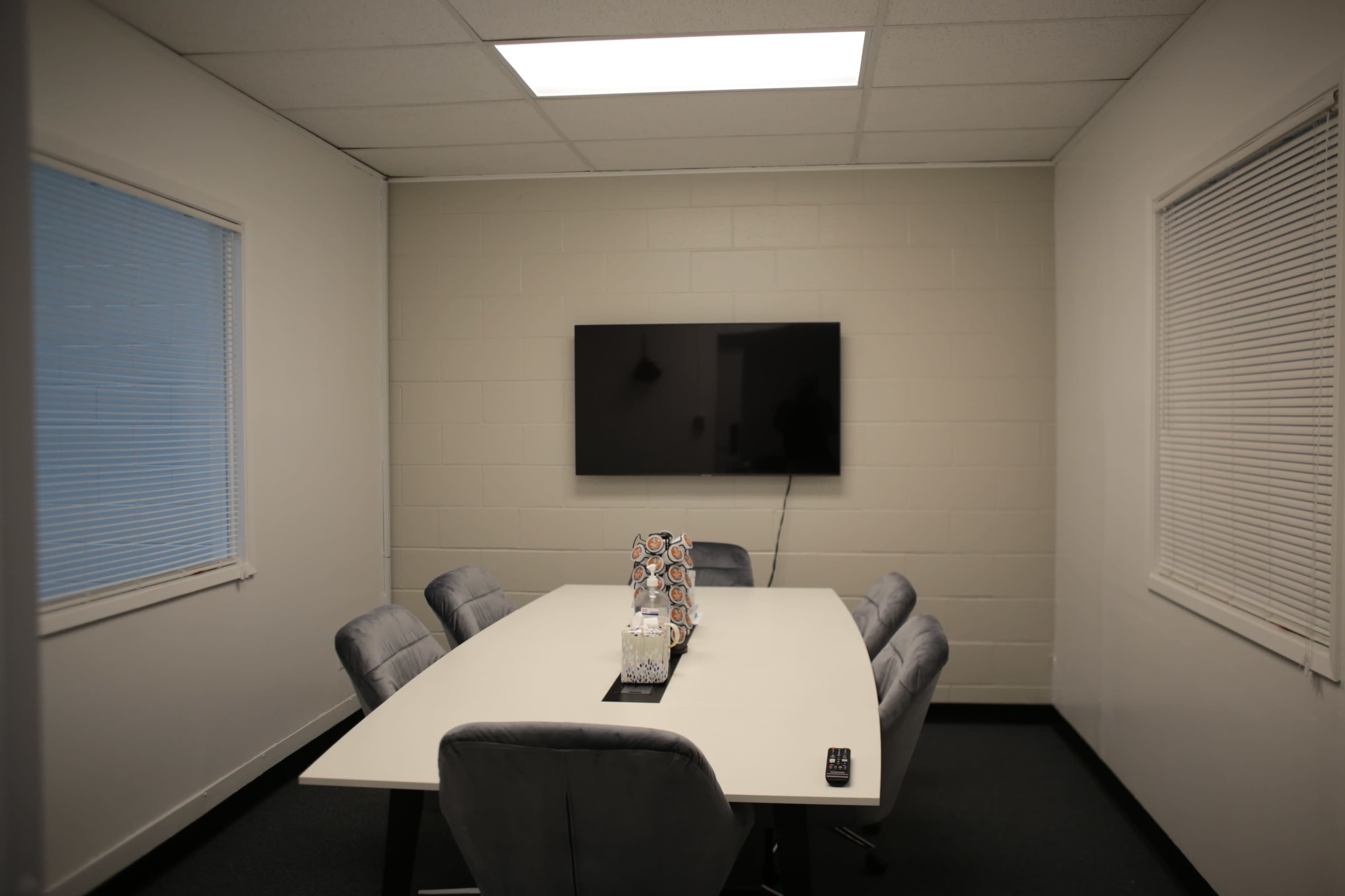 A conference room features a large table surrounded by six chairs, with a flat-screen TV mounted on the wall and two windows allowing natural light.