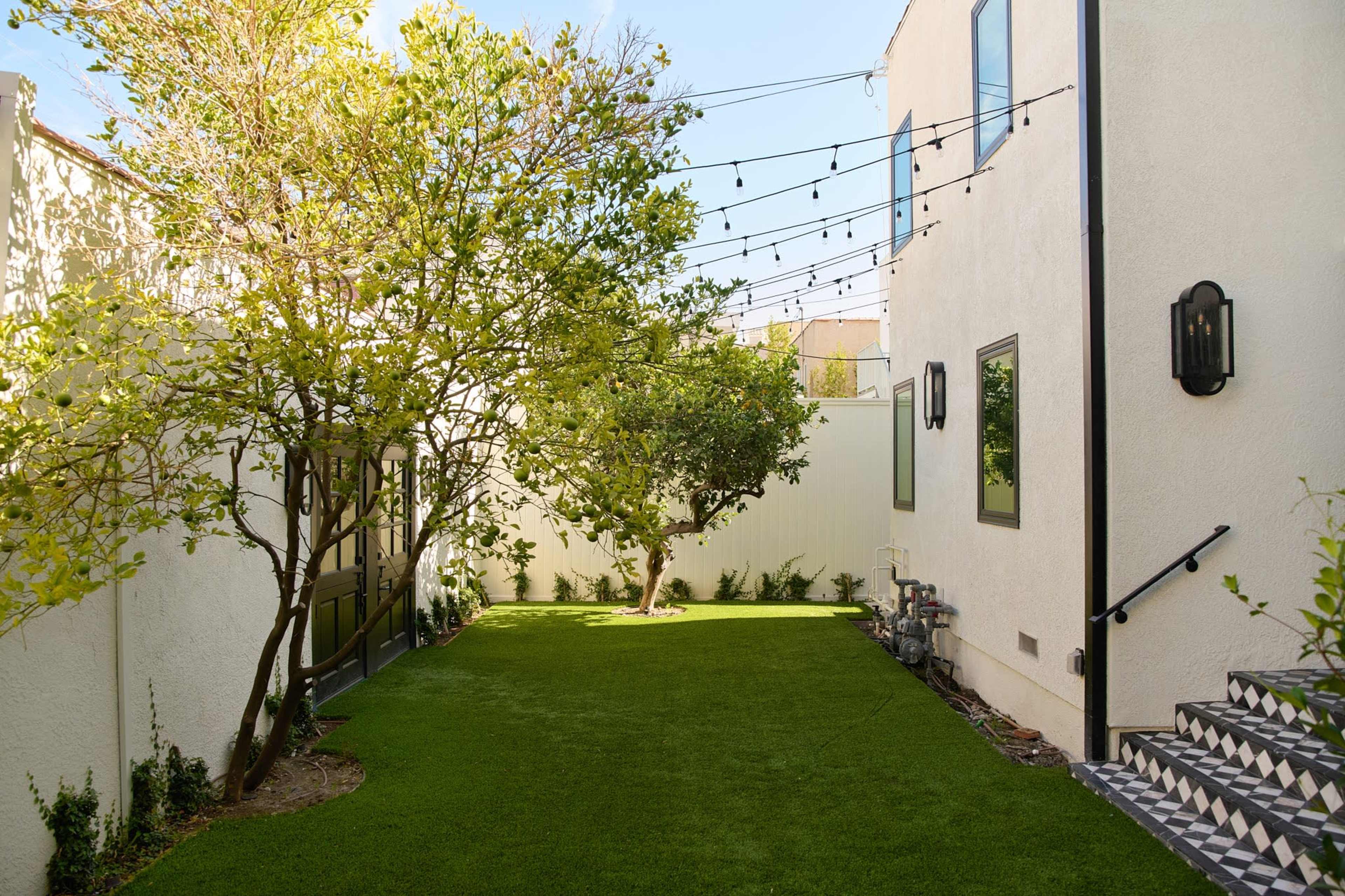 The image shows a landscaped backyard with artificial grass, a small tree, and a set of string lights overhead, framed by two white buildings.