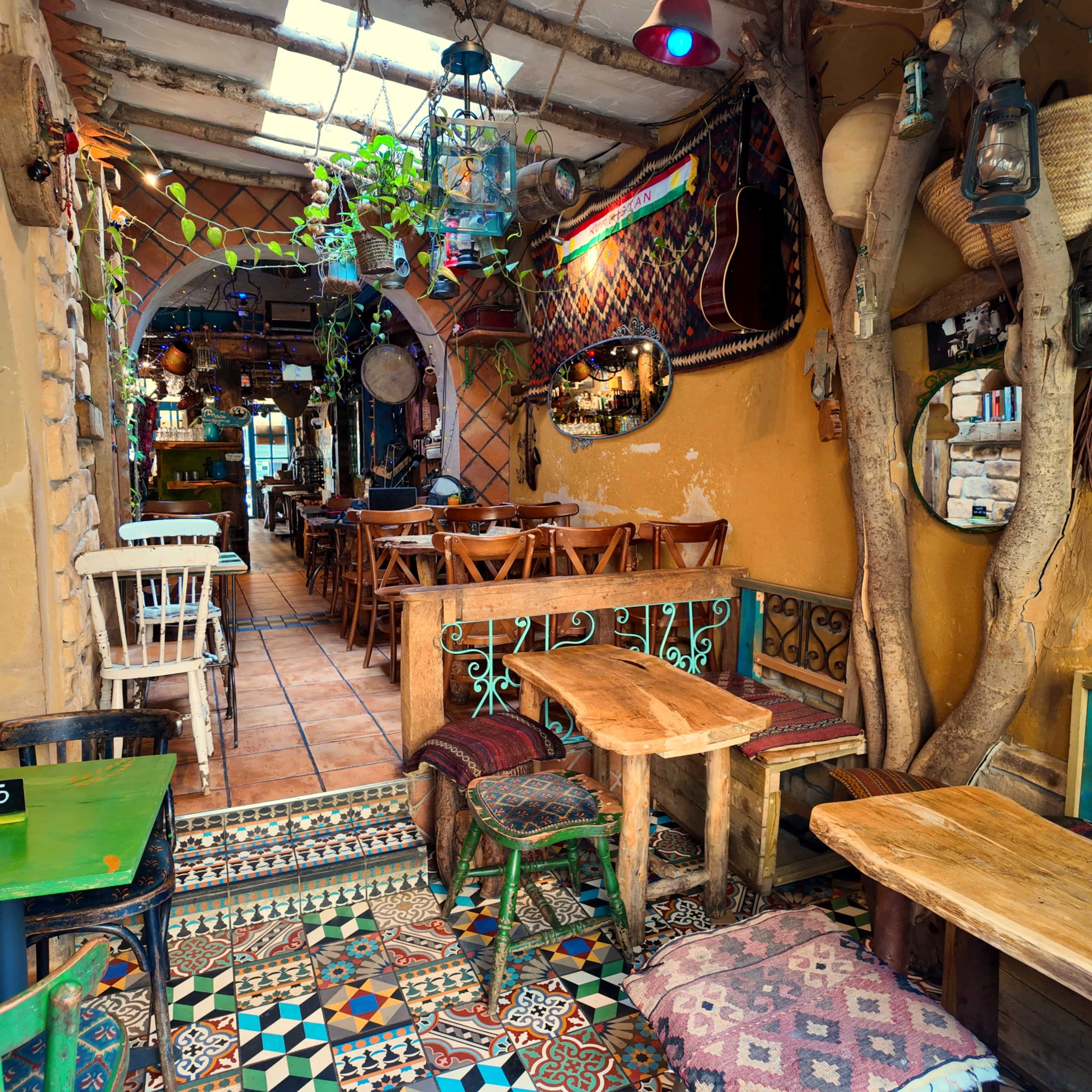 The image shows a vibrant café interior with wooden tables, colorful tiled flooring, and hanging plants, set against walls decorated with mirrors and artistic accents.