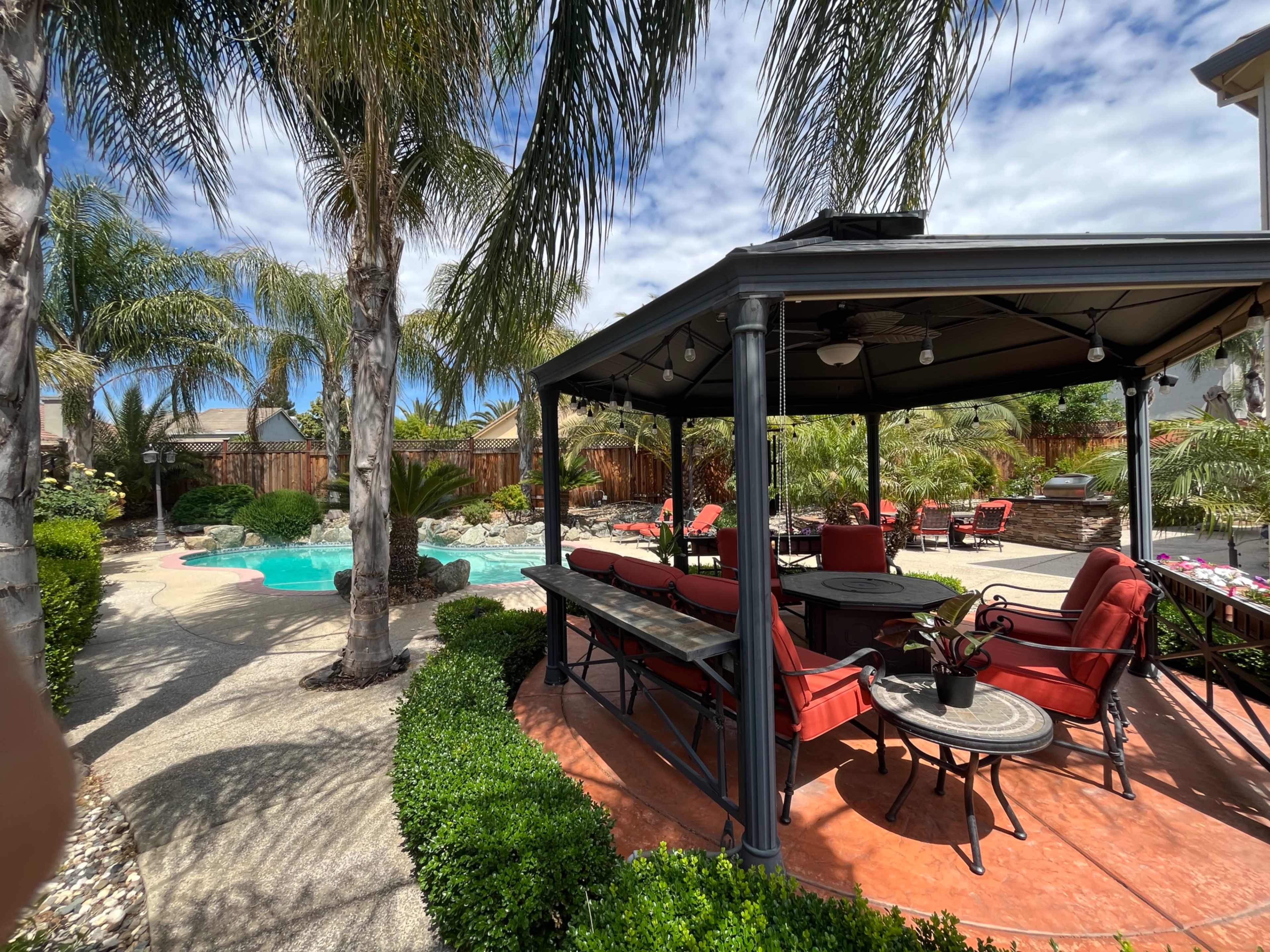A gazebo with seating surrounds a pool and is flanked by palm trees and landscaped gardens.