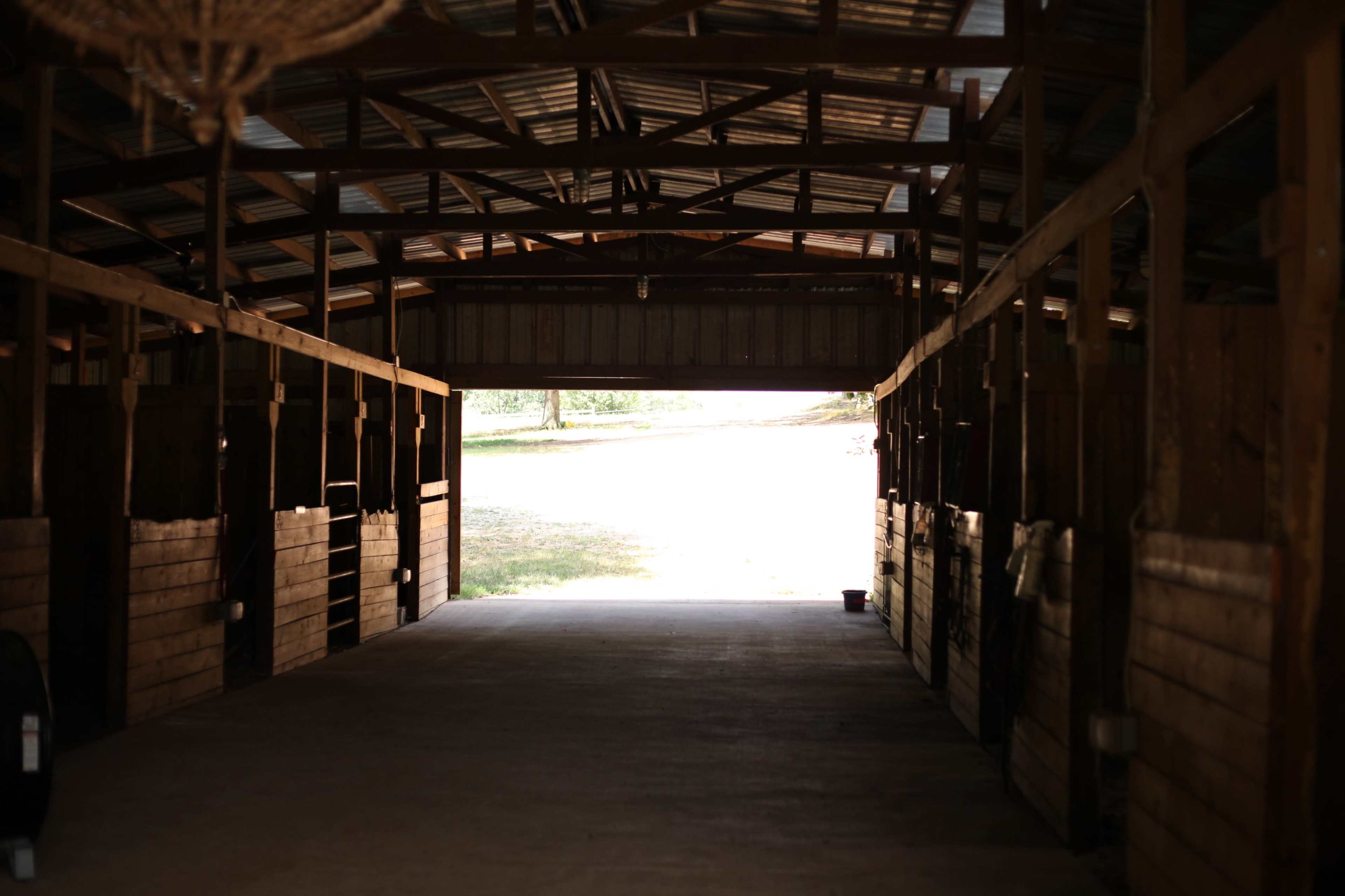 The image shows the interior of a barn with empty horse stalls lining the sides and light streaming in from an open doorway at the far end.